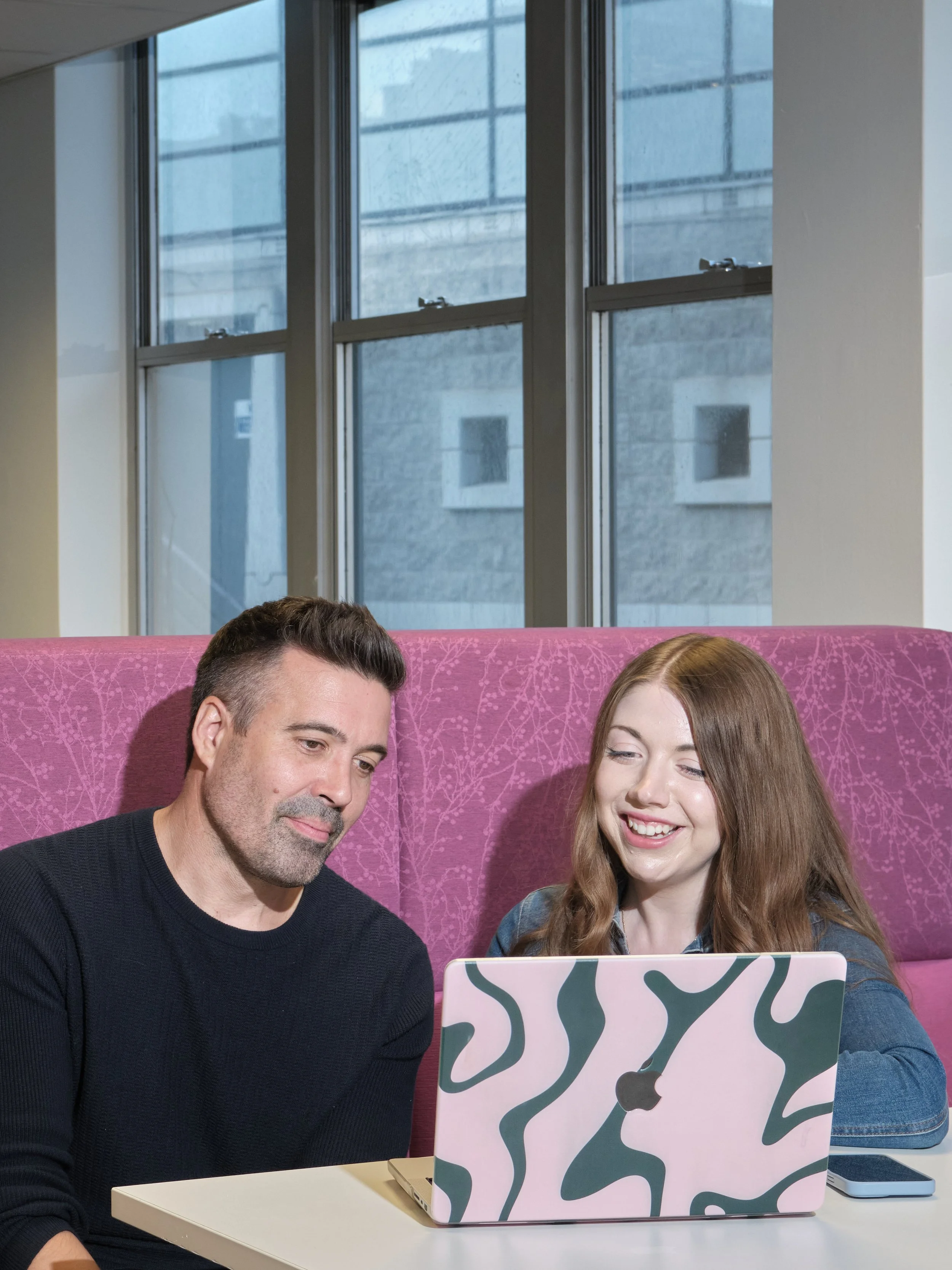 A man and a woman sitting at a white table, looking at a laptop with a pink, green, black, and white patterned cover, in a room with large windows and an urban view.