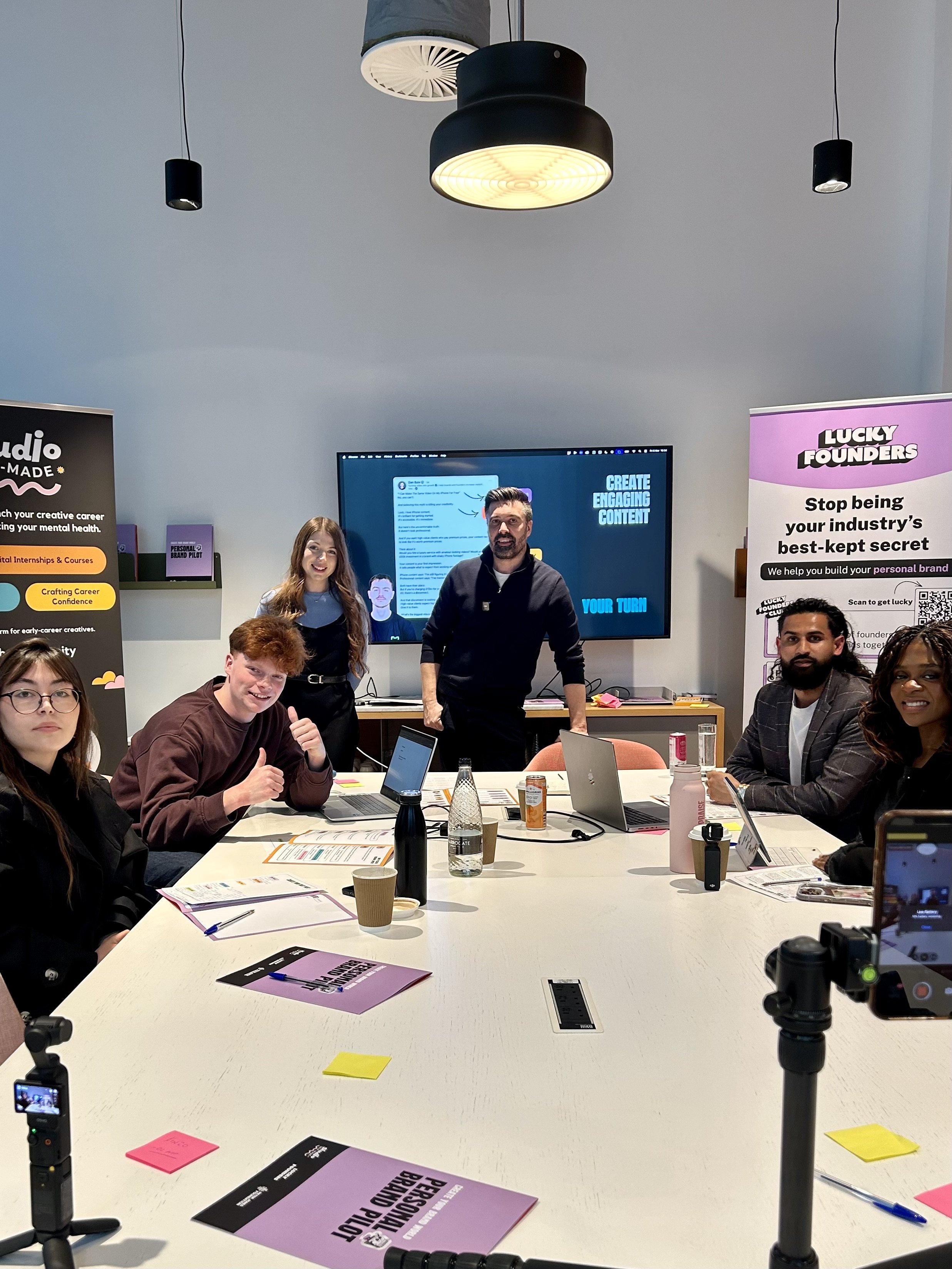 People seated around a conference table during a meeting or workshop in an office setting, with a man standing at the center, a woman smiling near him, and others seated at the table with laptops and documents.