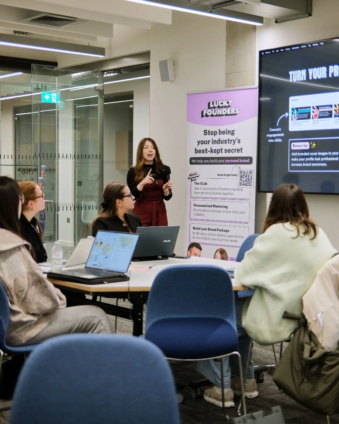 A woman giving a presentation to a group in a conference room, with a large screen displaying slides and a promotional banner in the background.