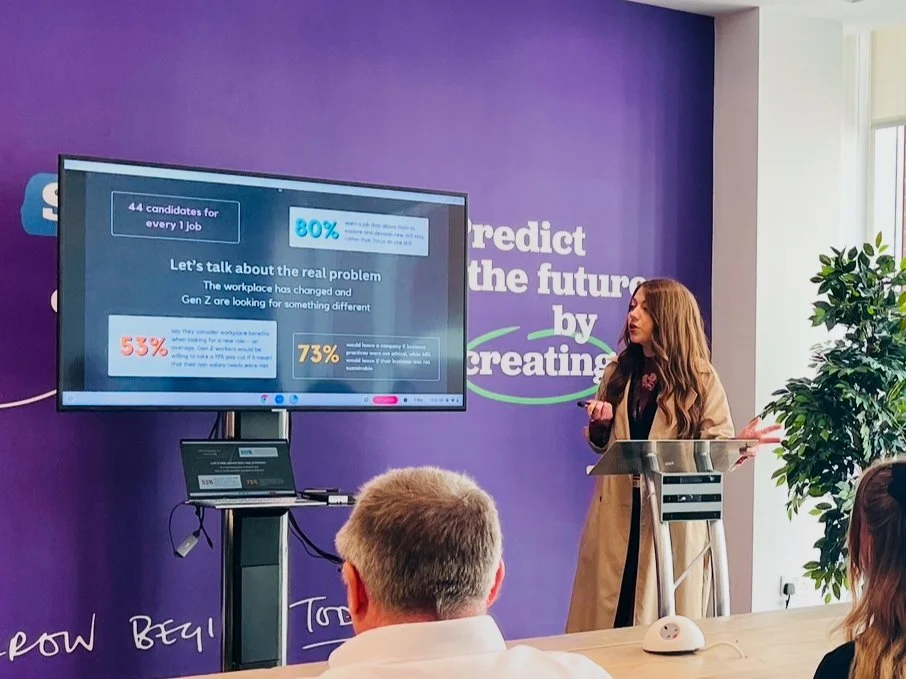 A woman gives a presentation in a conference room with a purple wall and a large screen displaying a slide about workplace change and employee needs. Three people are in the audience, listening attentively.