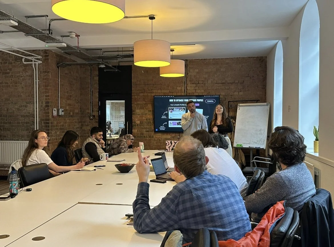 Business meeting in a conference room with a group of diverse people listening to two presenters at the front. There is a large screen displaying a presentation and a whiteboard on the side. The room has exposed brick walls and large windows with natural light.