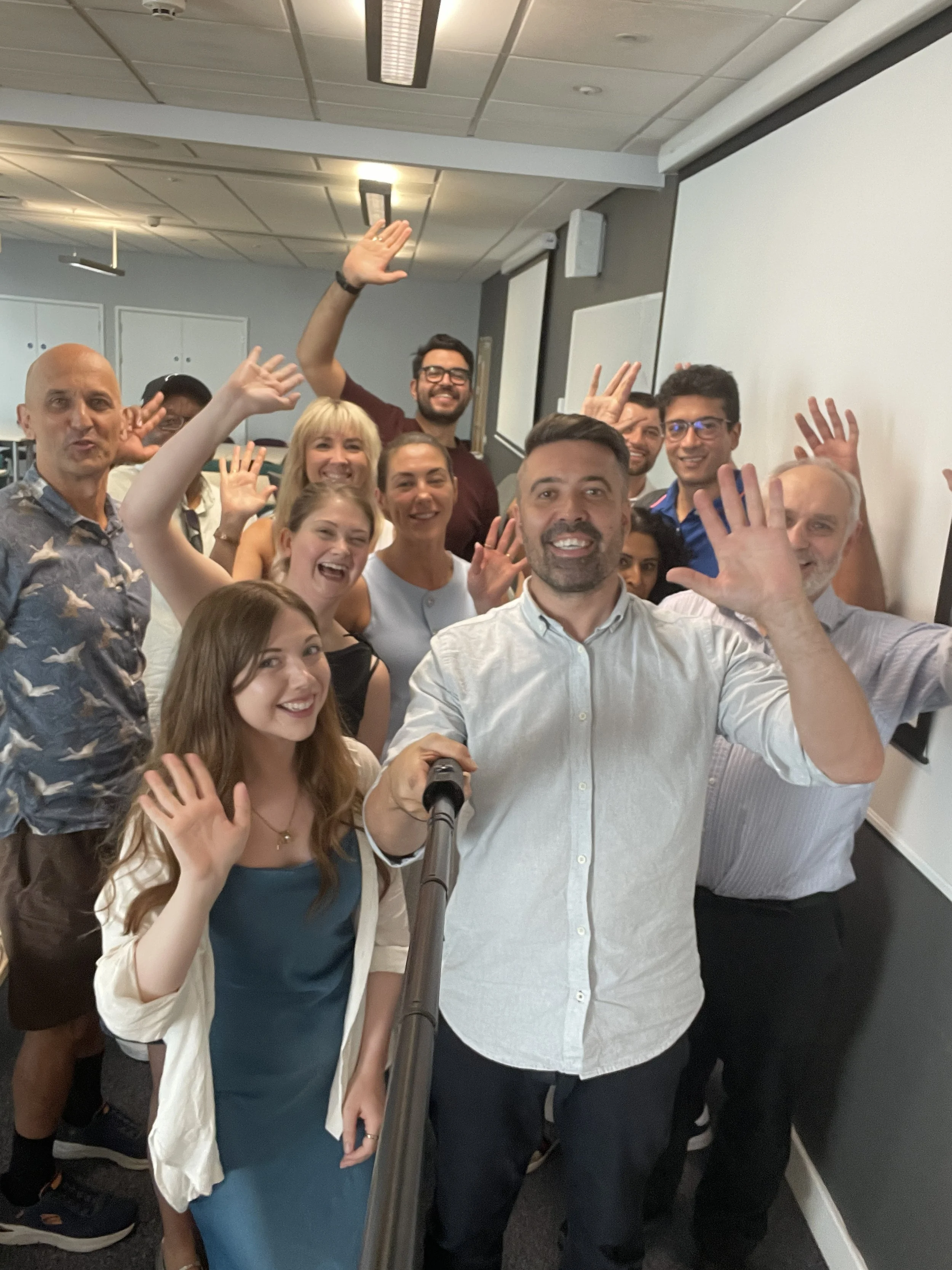 A group of diverse people, smiling and waving, taking a selfie in a conference room with a whiteboard, ceiling lights, and an air conditioning unit.