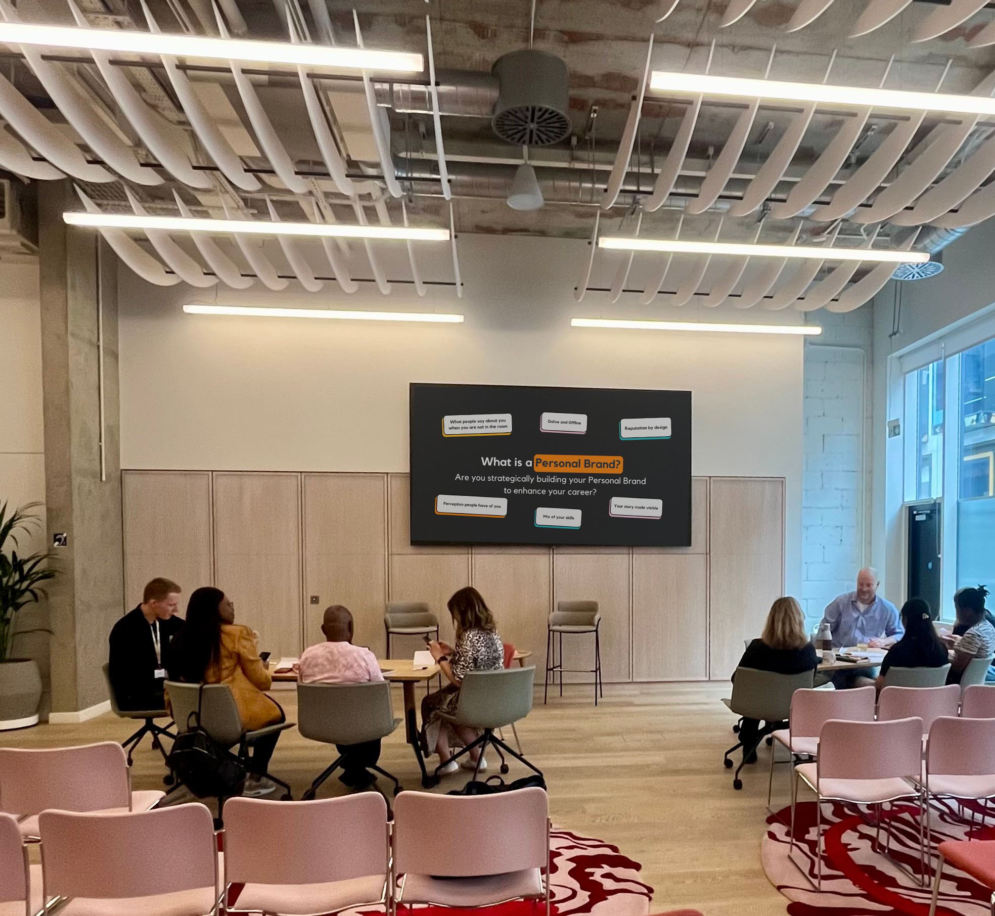 A group of people seated around tables in a modern conference room with pink chairs and a large screen displaying a presentation on personal branding.