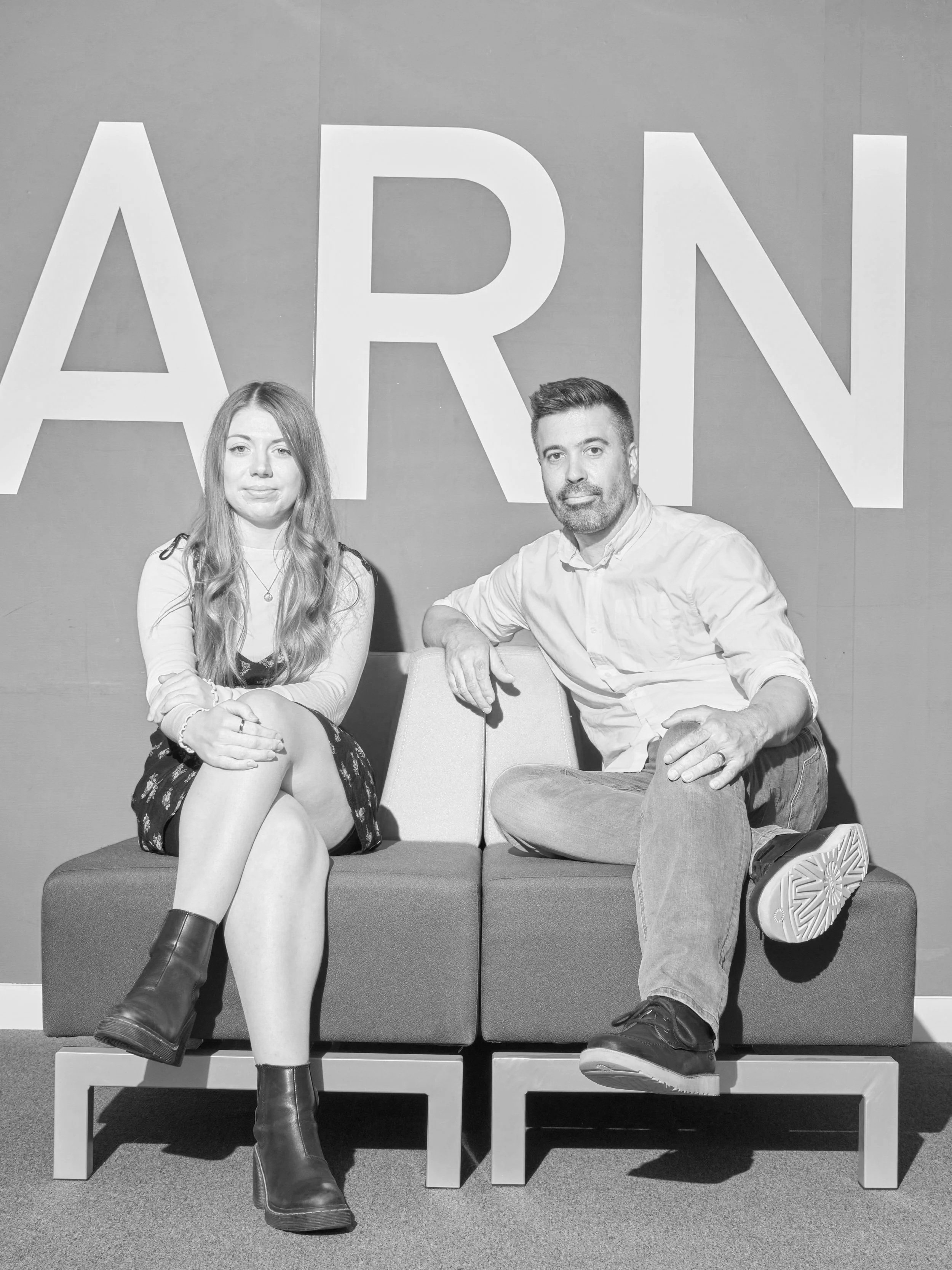 A woman and a man sitting on a bench in front of a wall with the letters 'ARN' displayed behind them. The woman has long hair, wears a dress with boots, and the man has short hair, a beard, wears a light-colored shirt and jeans.