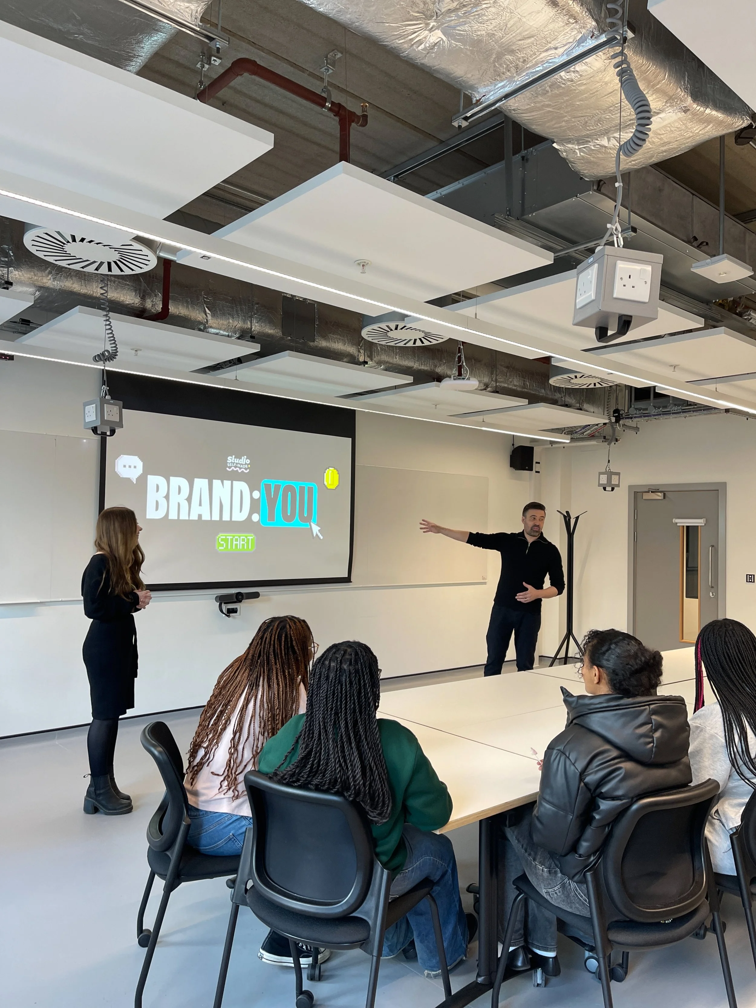 A classroom or conference room with a group of people seated around white tables, listening to a man standing and gesturing in front of a large screen that displays the words 'BRAND: YOU' with colorful text and graphics. The room has industrial ceiling design with white acoustic panels, vent covers, and electrical wiring visible.