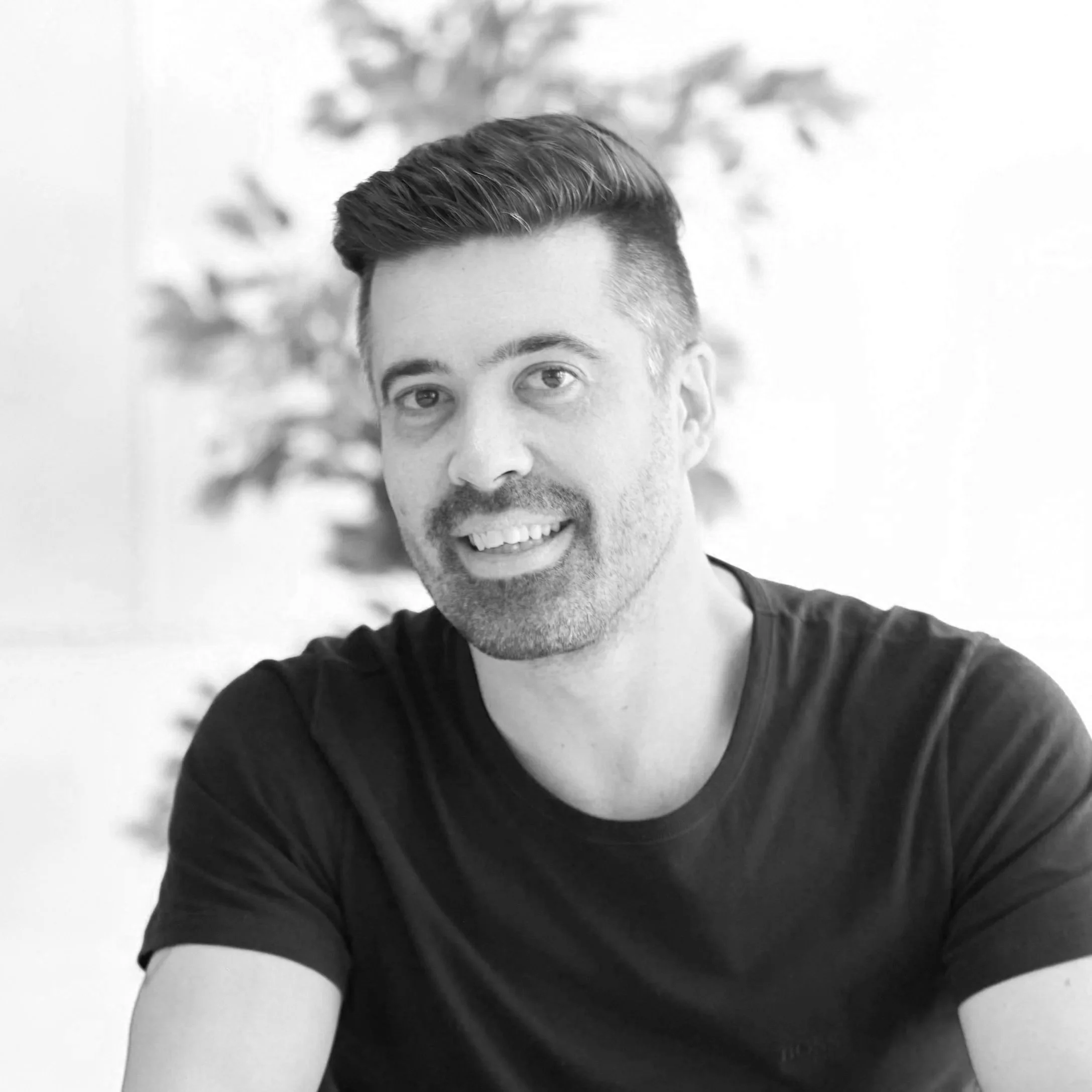 A black-and-white photo of a man smiling, with dark hair, facial hair, wearing a dark t-shirt, sitting indoors with a blurred plant in the background.