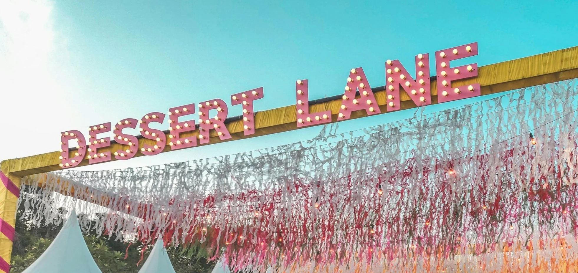 Pink marquee sign spelling 'DESSERT LANE' with white and pink hanging paper decorations underneath against a clear blue sky.