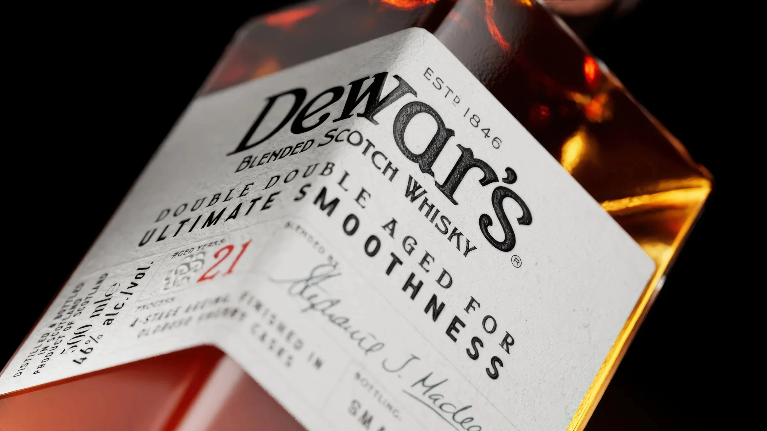 Close-up of a bottle of Dewar's Blended Scotch Whisky against a black background, showing the label with black and red text.