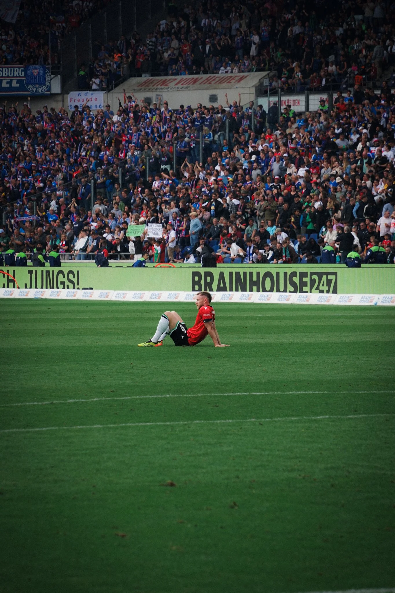 Fußballspieler sitzt auf dem Rasen während eines Spiels, mit vollen Tribünen im Hintergrund.