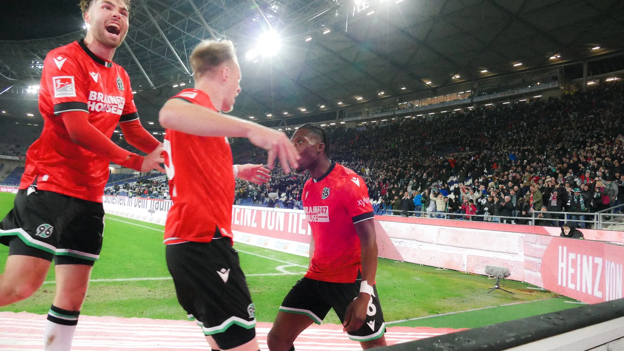 Fußballspieler im roten Trikot mit der Nummer 96 feiern einen Erfolg auf dem Spielfeld, während das Publikum im Hintergrund jubelt.