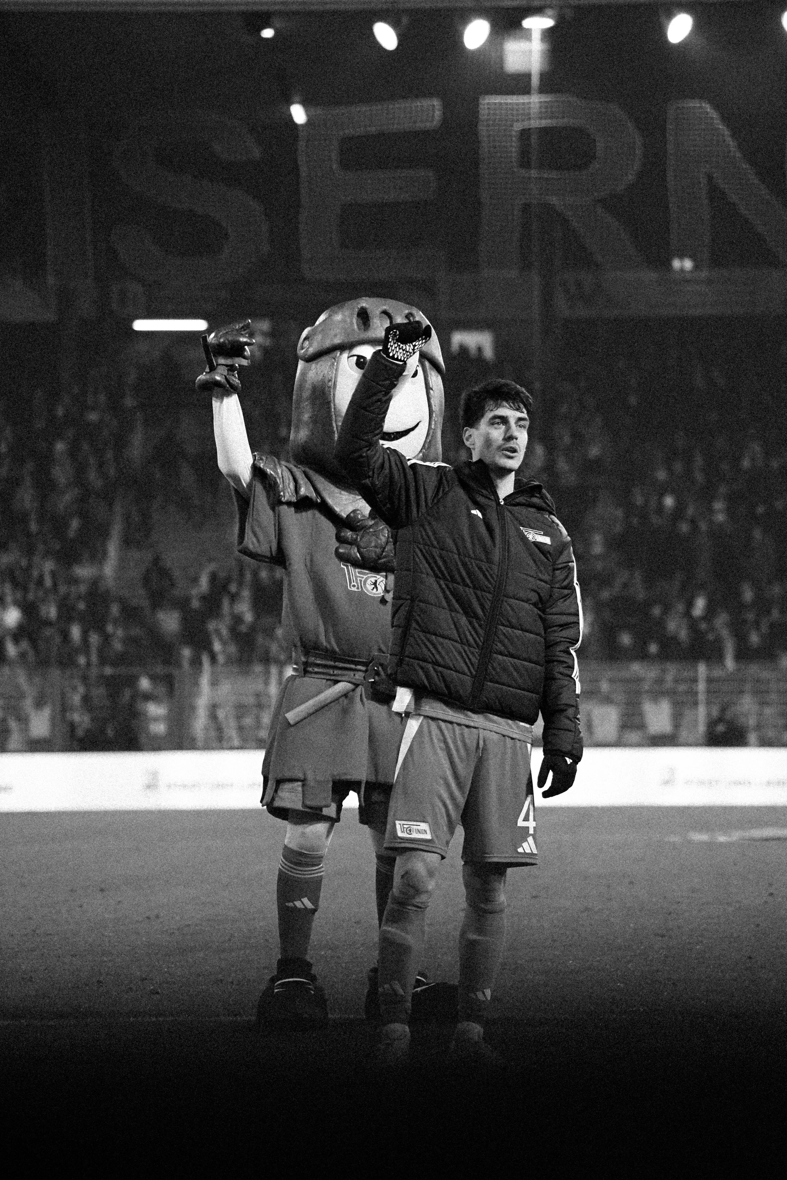 Ein Fußballspieler und ein Maskottchen stehen auf einem Spielfeld. Der Spieler trägt eine Trainingsjacke und Handschuhe, während das Maskottchen in einem Ritterkostüm gekleidet ist. Beide heben ihre Hände in die Luft, um den Sieg zu feiern. Im Hinter