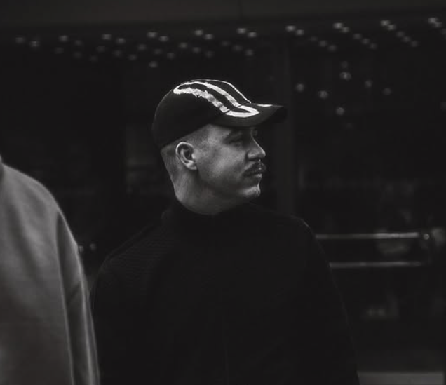 Schwarz-weiß-Foto eines Mannes mit Basecap, der nach rechts schaut, vor dunklem Hintergrund.