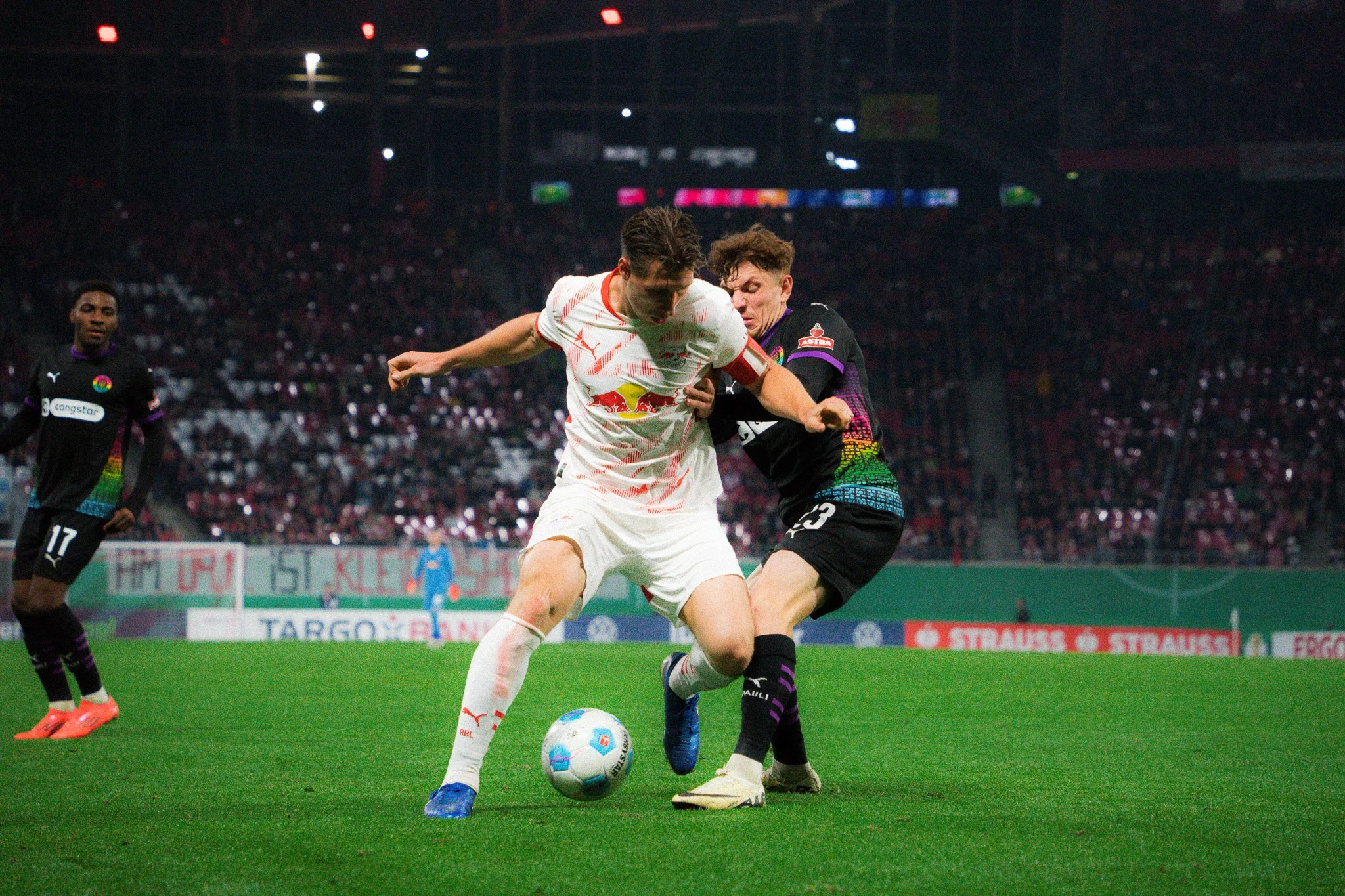 Fußballspieler im Zweikampf während eines Spiels. Der Spieler in Weiß trägt das Trikot von RB Leipzig, während der Gegenspieler ein schwarzes Trikot trägt. Der Ball ist zwischen ihnen. Im Hintergrund sind leere Ränge eines Stadions zu sehen.