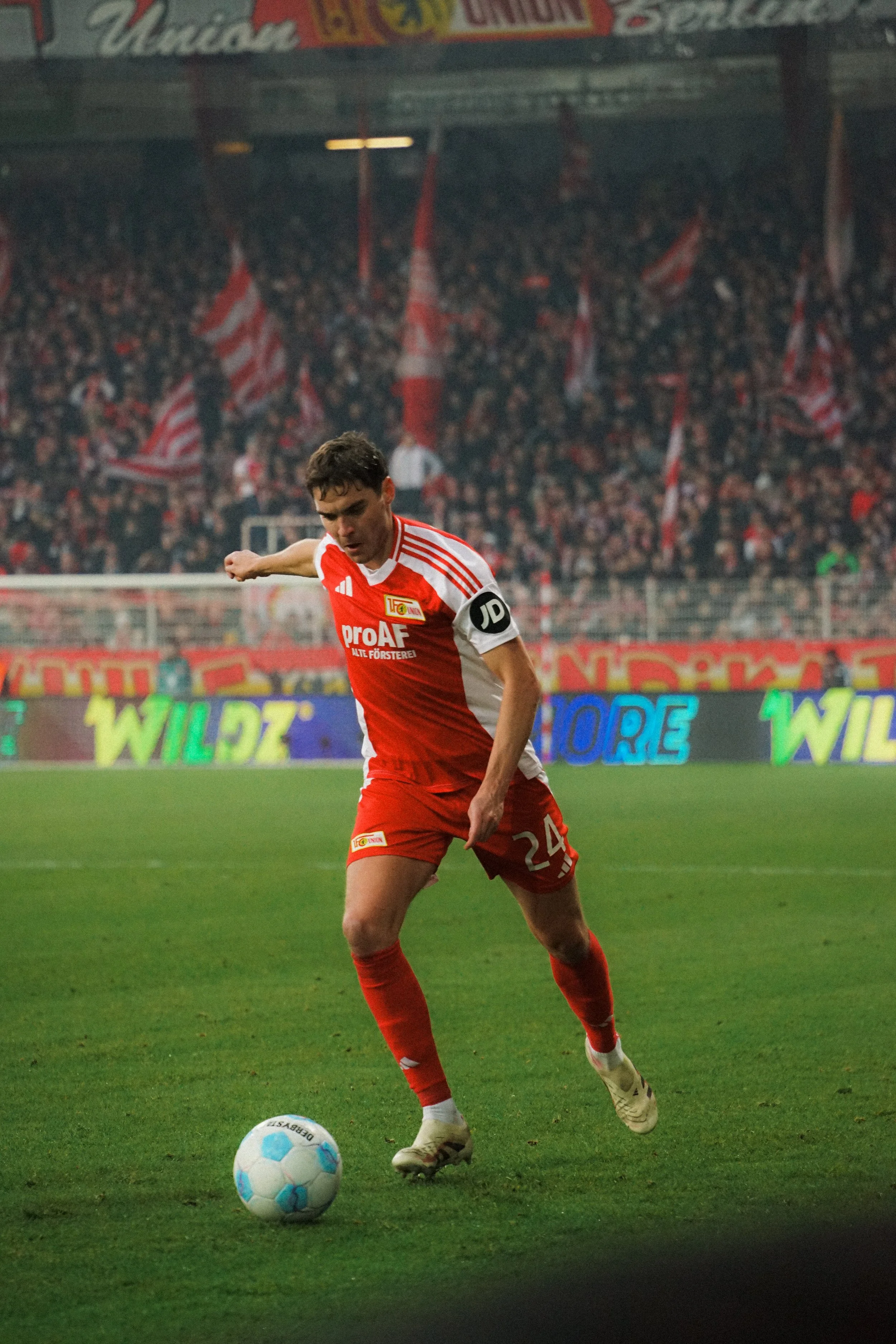 Ein Fußballspieler in einem roten Trikot läuft auf dem Spielfeld in einem Stadion. Im Hintergrund sind Fans mit roten Fahnen zu sehen.