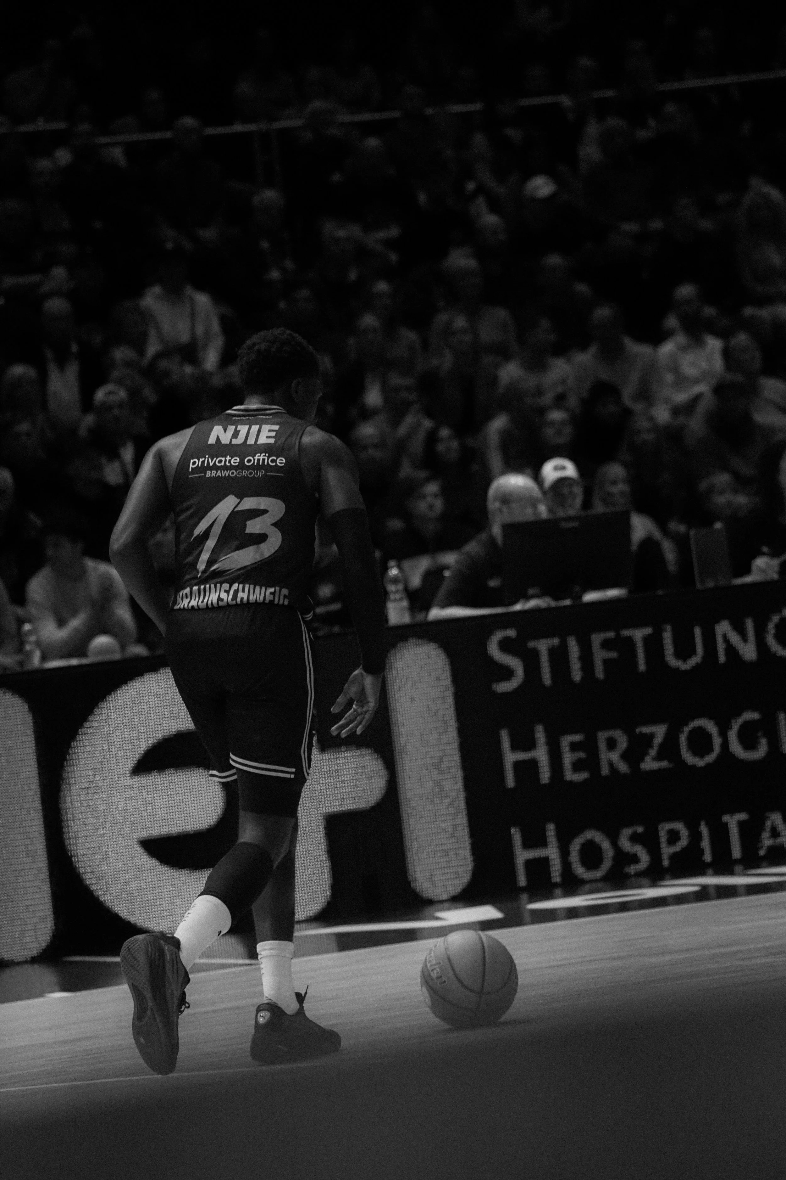 Ein Basketballspieler in einem dunklen Trikot mit der Nummer 13 dribbelt den Ball auf einem Spielfeld. Im Hintergrund sind Zuschauer und Werbebanner zu sehen.