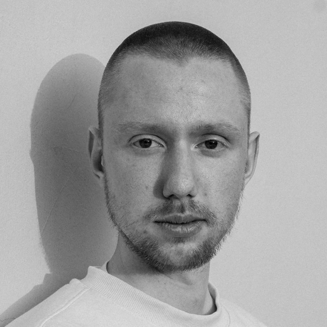 Schwarz-weiß Foto eines Mannes mit kurzem Haarschnitt und Bart, der direkt in die Kamera schaut, vor einer neutralen Hintergrundwand.