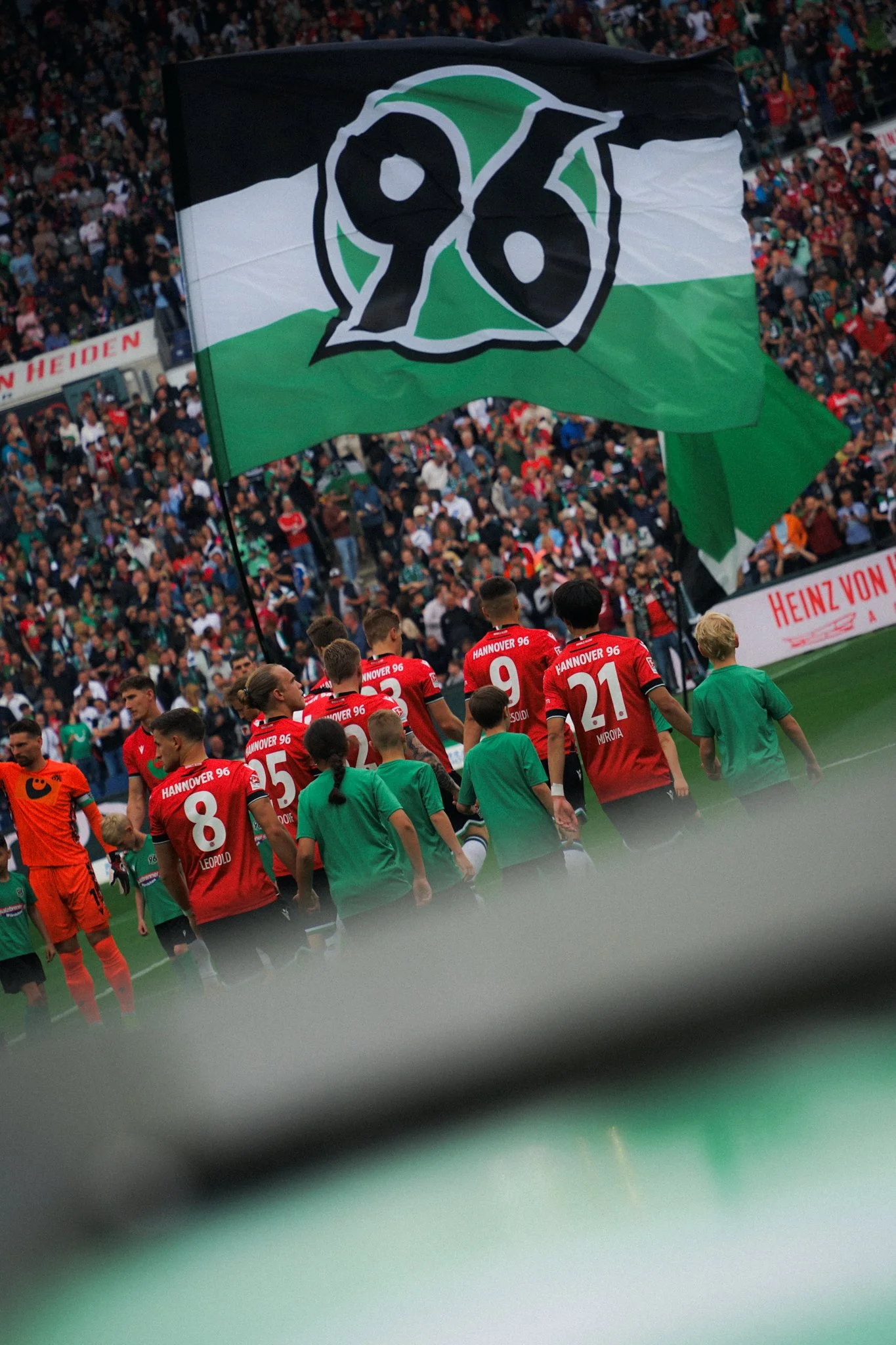 Fußballspieler von Hannover 96 auf dem Spielfeld mit einer großen Flagge im Hintergrund und Zuschauern im Stadion.
