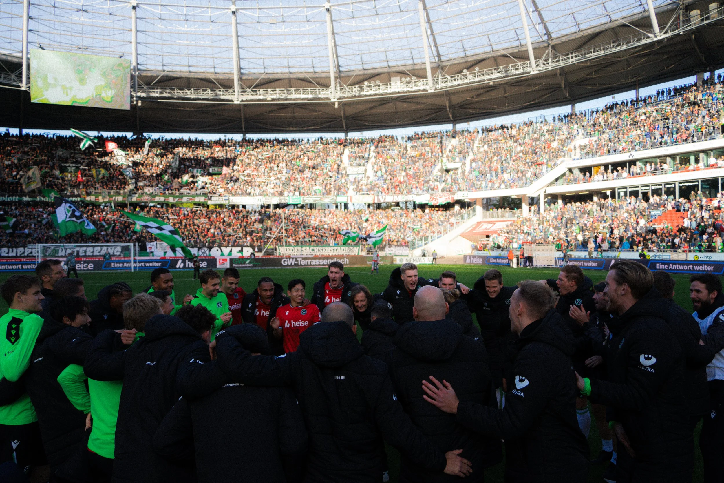 Fußballspieler und Trainer im Stadion versammeln sich im Kreis, während das Publikum im Hintergrund jubelt.