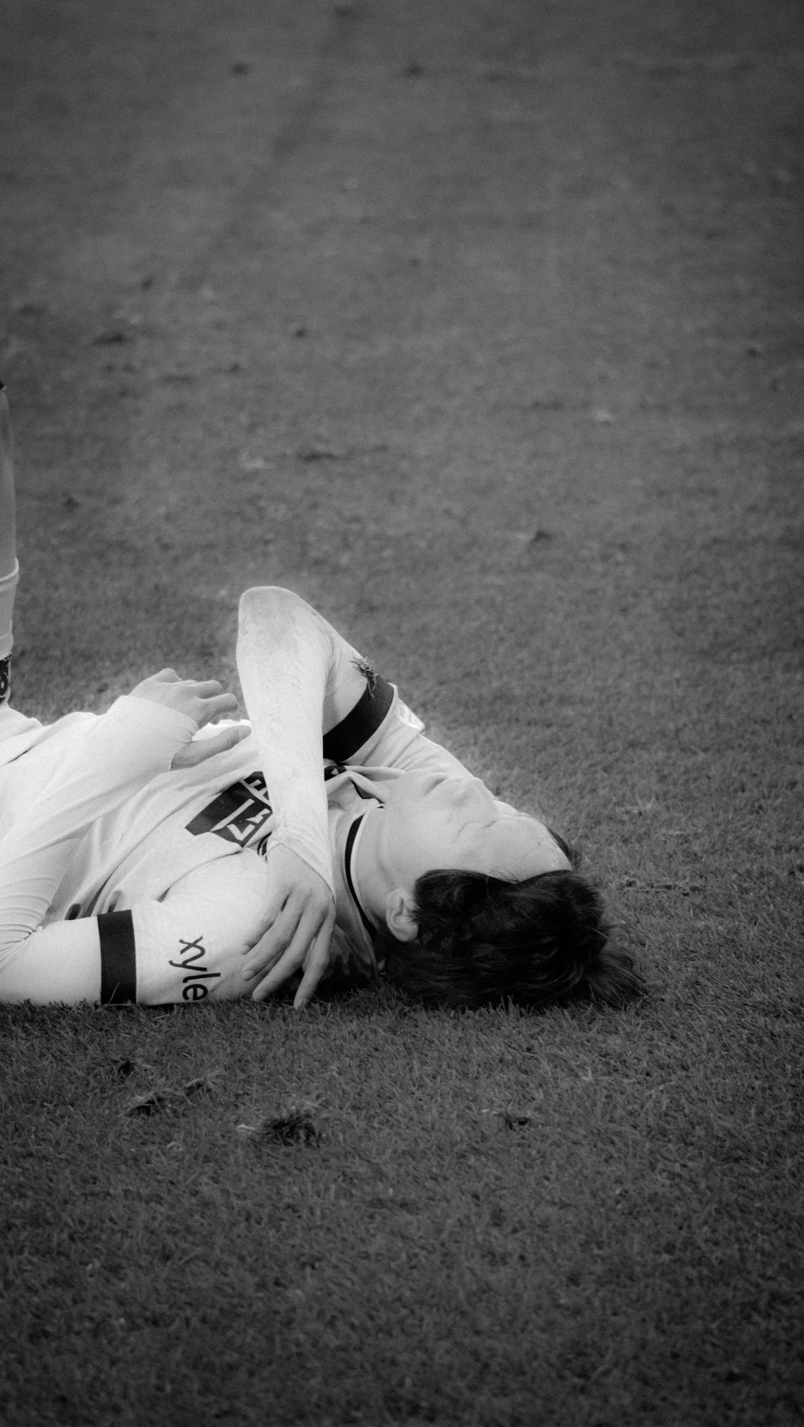 Schwarzweißbild eines Fußballspielers, der verletzt am Boden liegt. Der Spieler trägt ein Trikot und hält sich den Arm. Im Hintergrund ist Rasengras zu sehen.