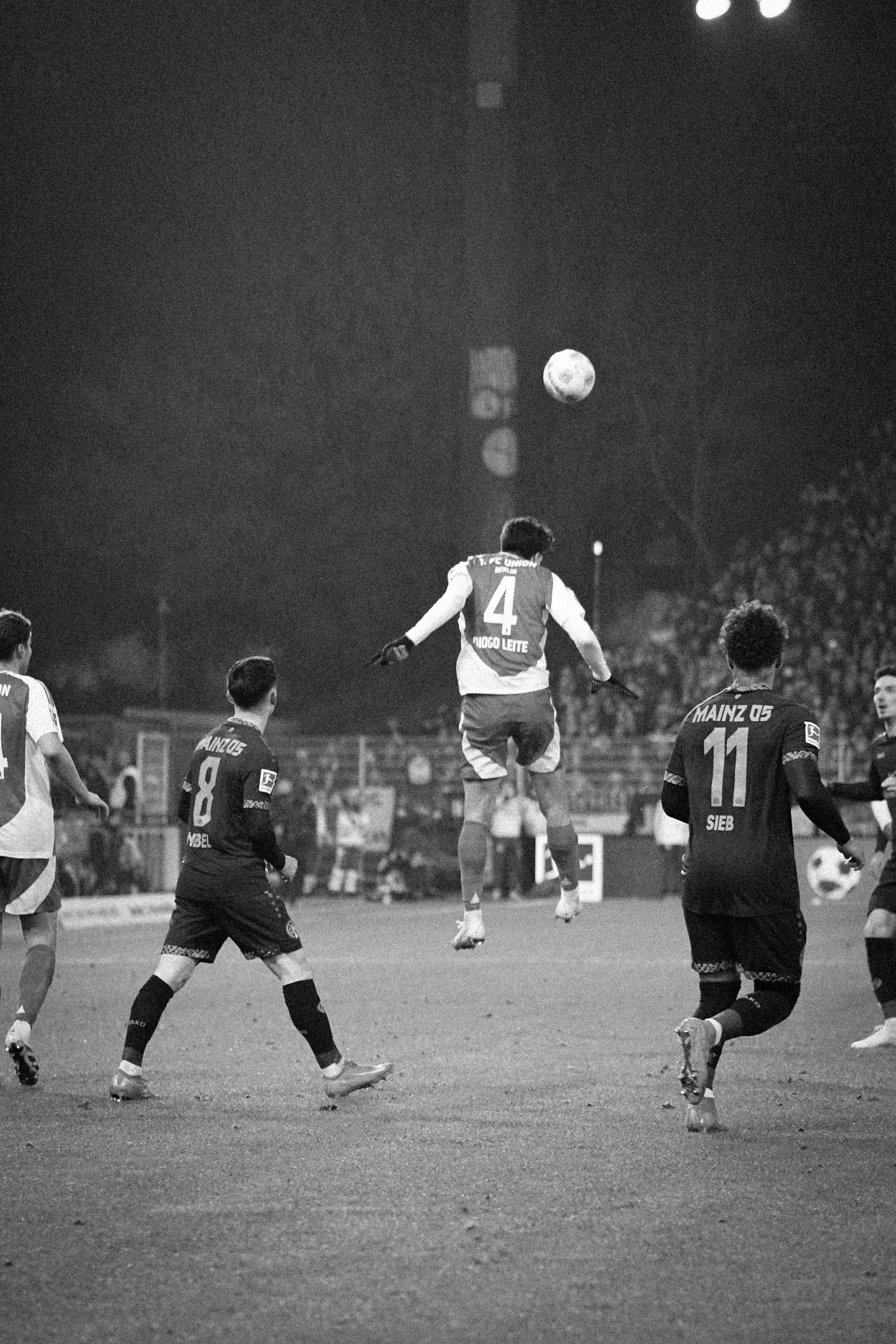 Fußballspiel bei Nacht; Spieler in Aktion, einer springt zum Kopfball; Trikots tragen Markierungen "FC UNION" und "MAINZ 05"; schwarz-weiße Fotografie.