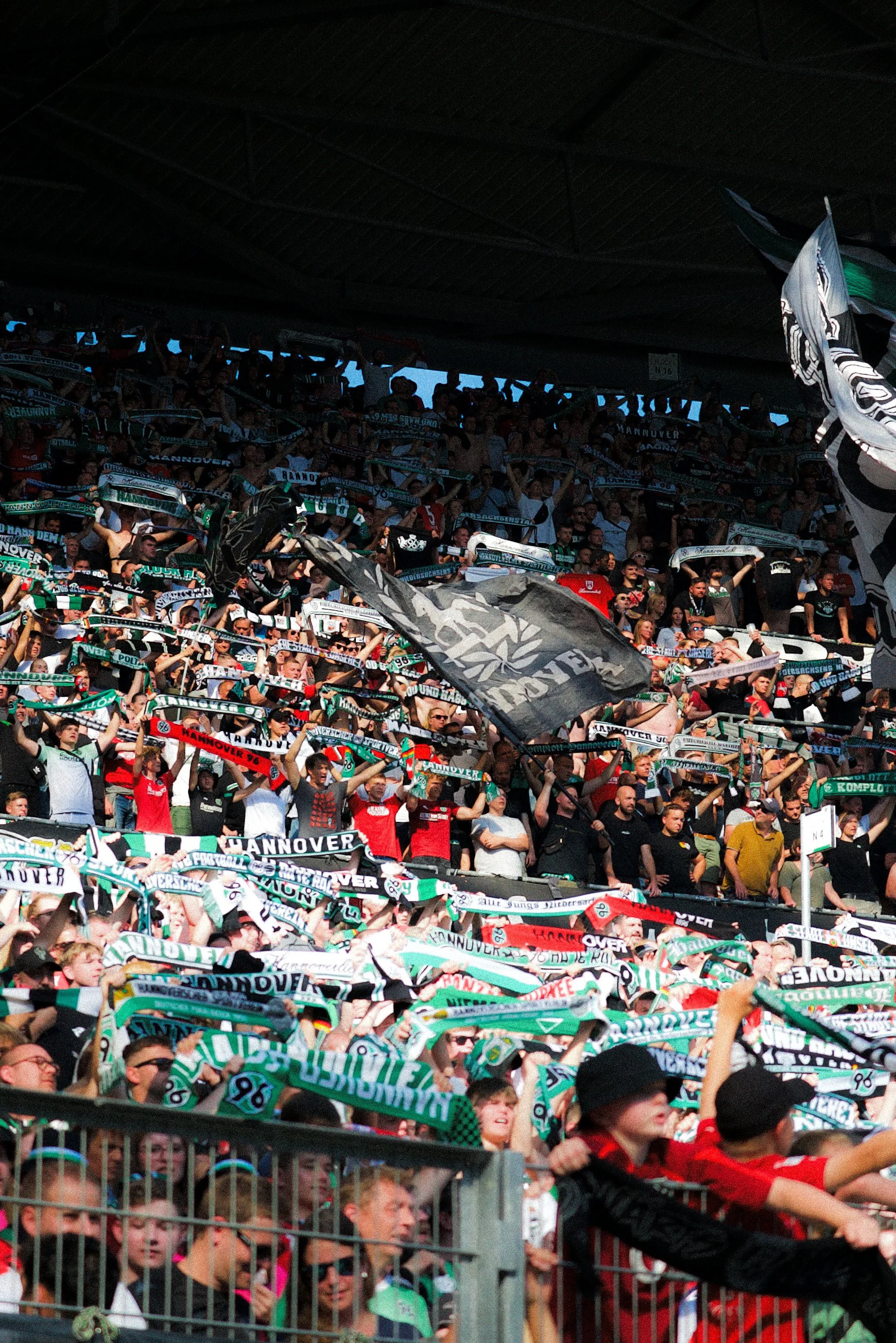 Fußballfans im Stadion, die Schals hochhalten und jubeln, viele tragen Rot und Grün, einige schwenken Fahnen.