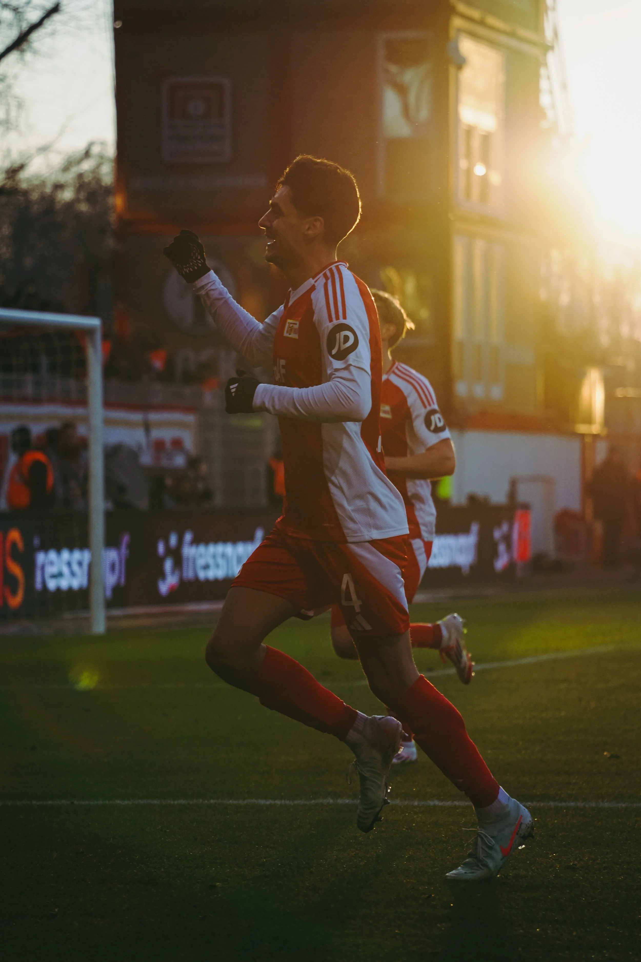 Ein Fußballspieler mit rotem und weißem Trikot jubelt auf dem Spielfeld bei Sonnenuntergang. Im Hintergrund sind Zuschauer und eine Tribüne zu sehen.