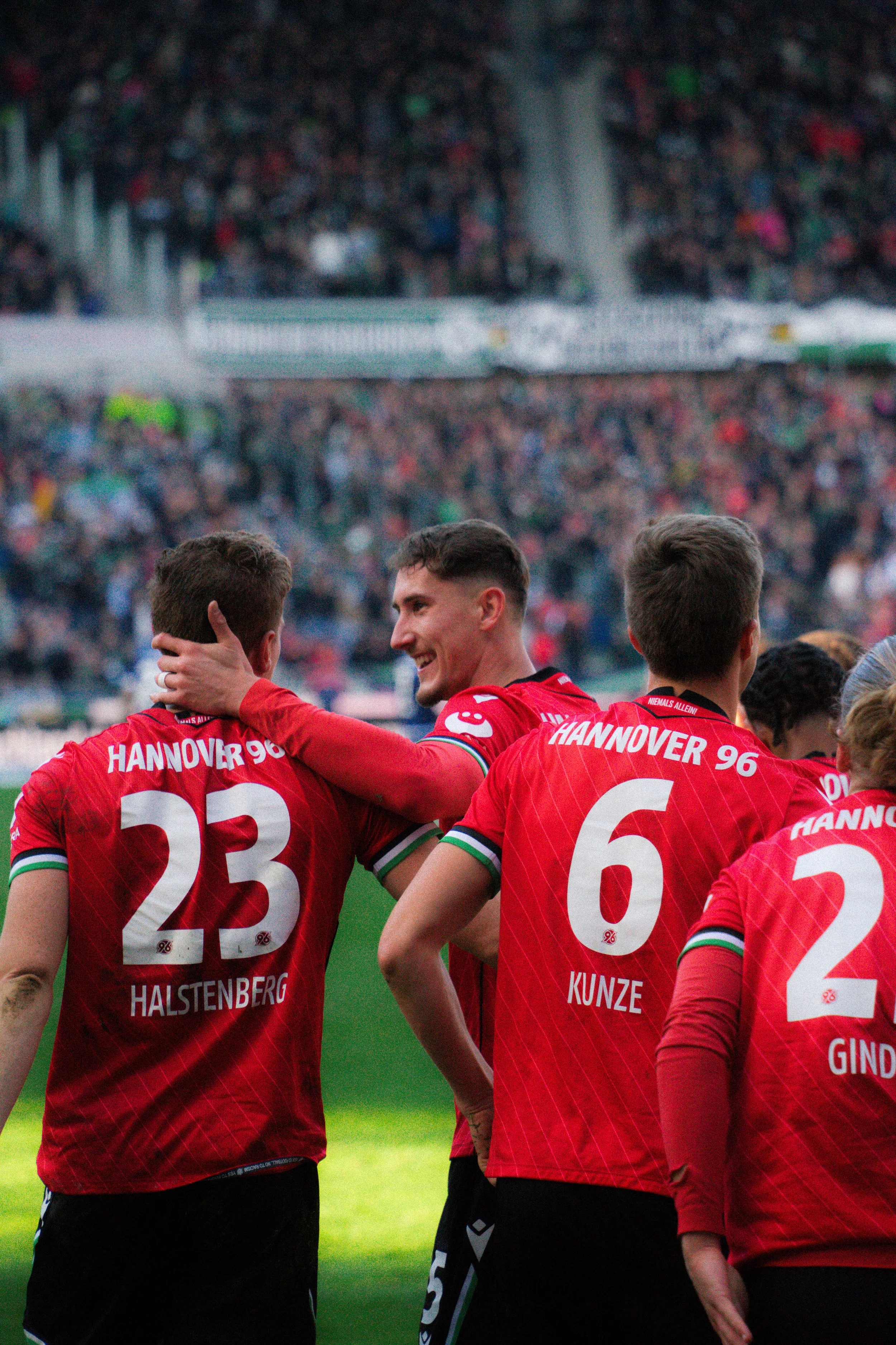 Fußballspieler von Hannover 96 feiern auf dem Spielfeld, gekleidet in roten Trikots mit den Namen und Nummern Halstenberg (23), Kunze (6) und Gindorf erkennbar.