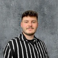 Porträt eines lächelnden Mannes mit dunklem Haar und Bart, der ein Hemd mit schwarzen und weißen Streifen trägt vor einem grauen Hintergrund.