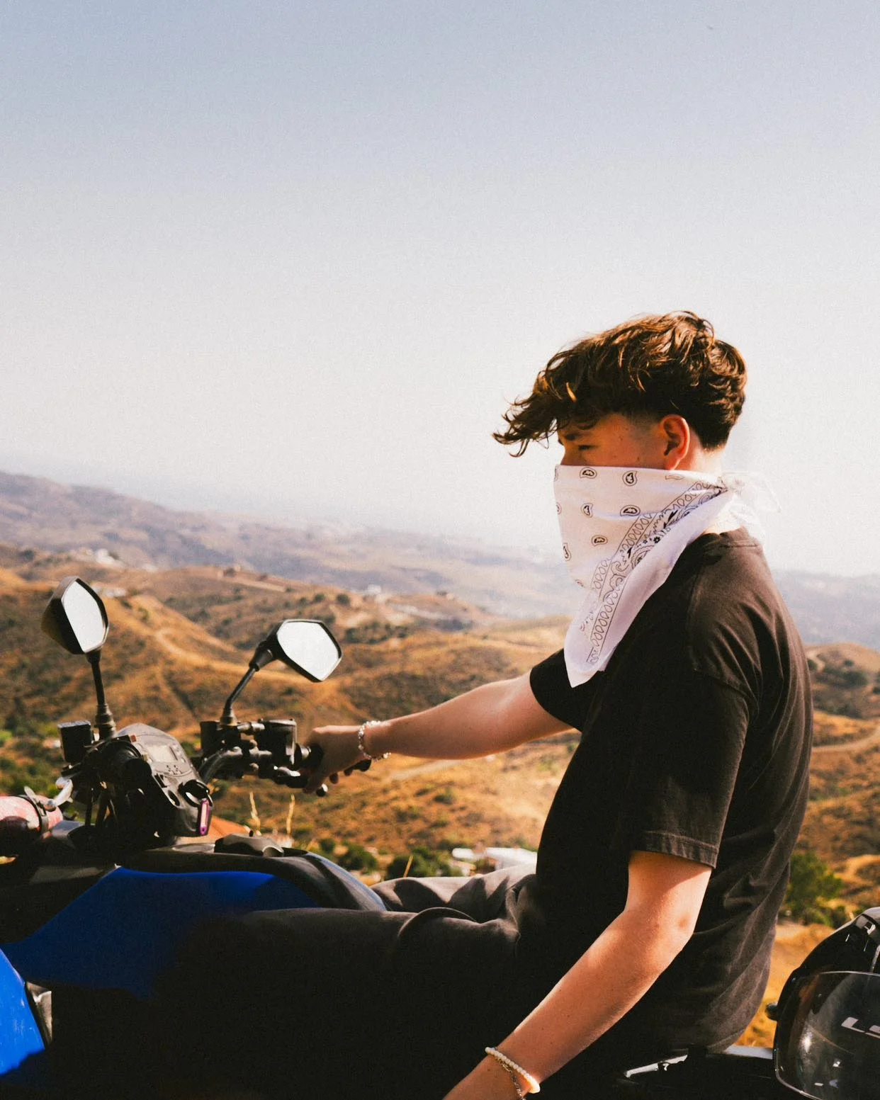 Ein Mensch auf einem Motorrad mit bergiger Landschaft im Hintergrund, trägt ein schwarzes T-Shirt und ein weißes Halstuch.
