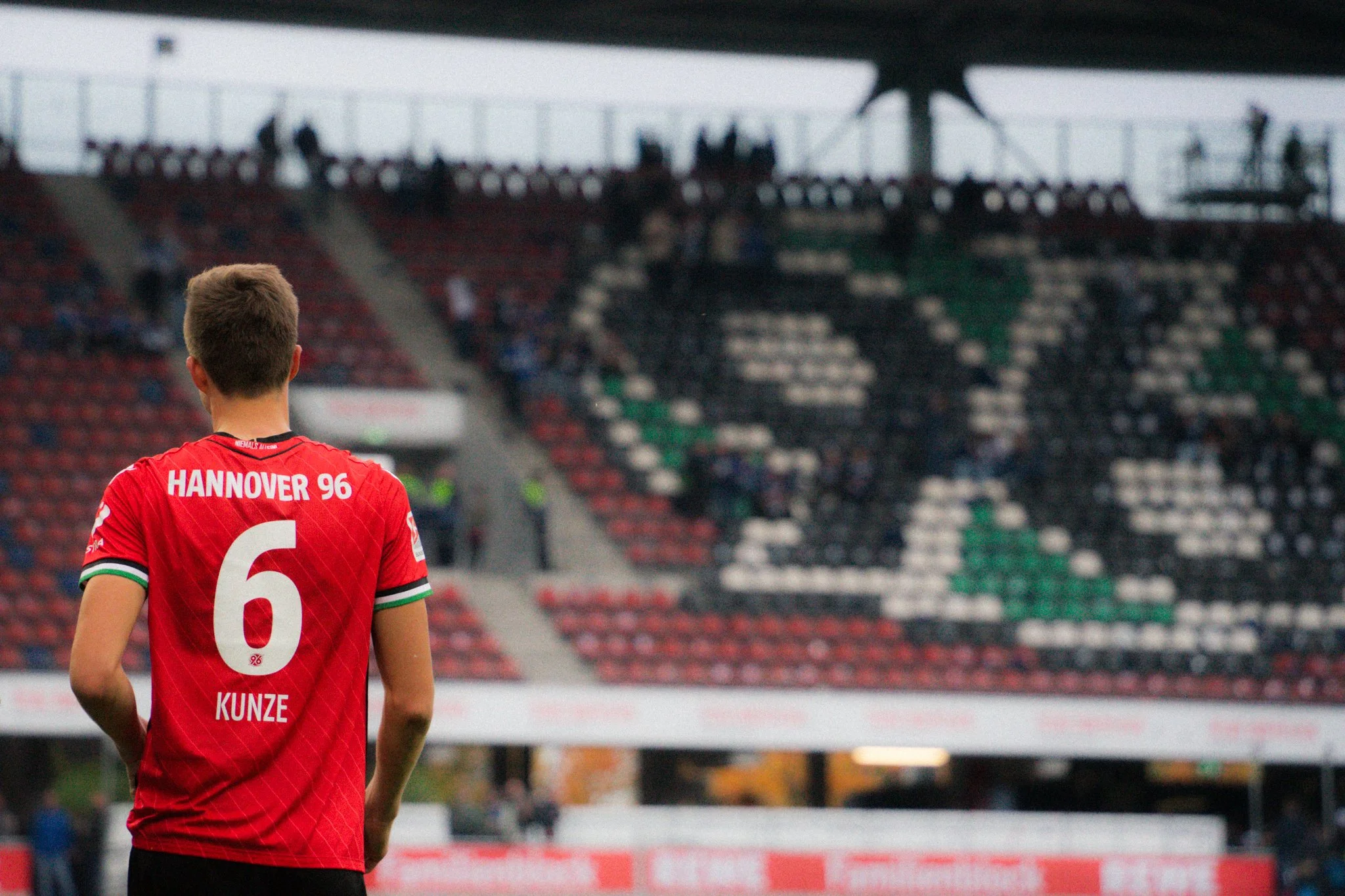 Ein Fußballspieler von hinten im roten Trikot von Hannover 96 mit der Nummer 6 und dem Namen Kunze. Im Hintergrund ist ein fast leeres Stadion zu sehen, das Logo von Hannover 96 auf den Sitzen erkennbar.