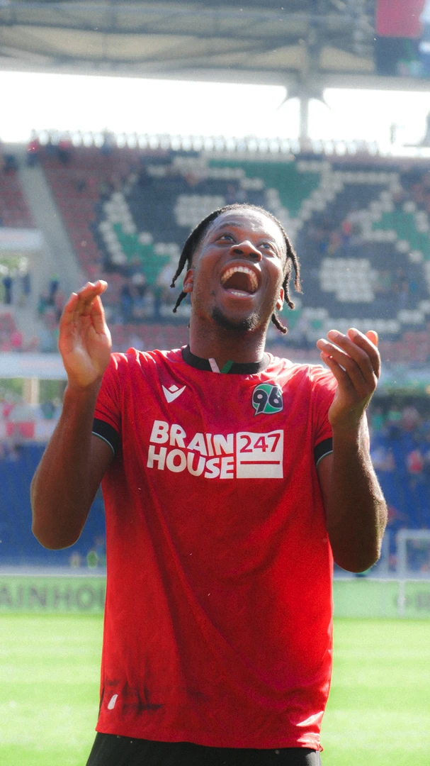 Ein Fußballspieler im roten Trikot von Hannover 96 erfreut sich auf dem Spielfeld. Im Hintergrund ist ein teilweise besetztes Stadion zu sehen.
