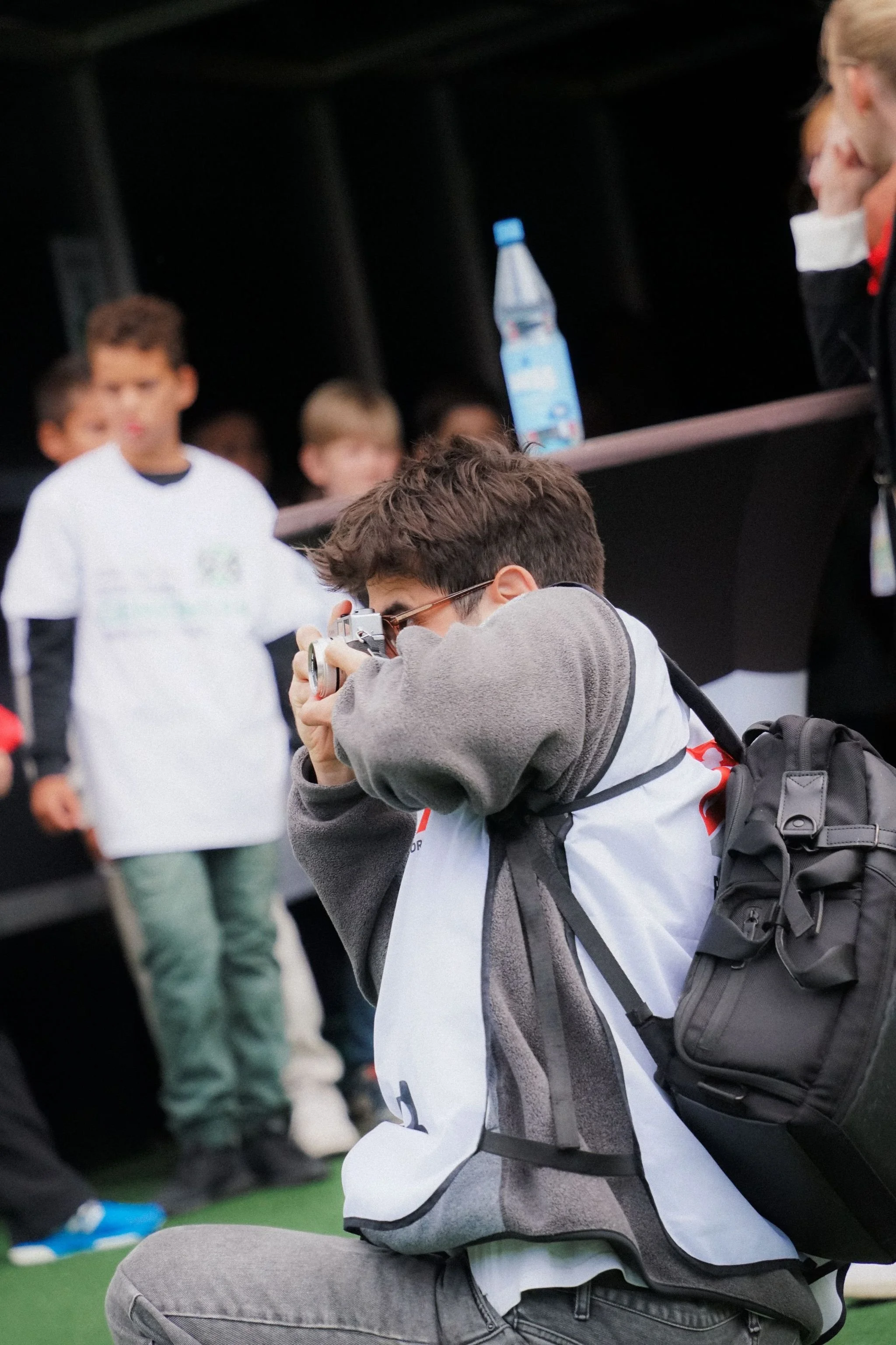 Ein Fotograf kniet und macht Fotos von einer Gruppe von Kindern, die im Hintergrund stehen. Er trägt eine Brille, ein graues Sweatshirt und hat einen schwarzen Rucksack auf dem Rücken. Auf einem Tisch steht eine Wasserflasche.