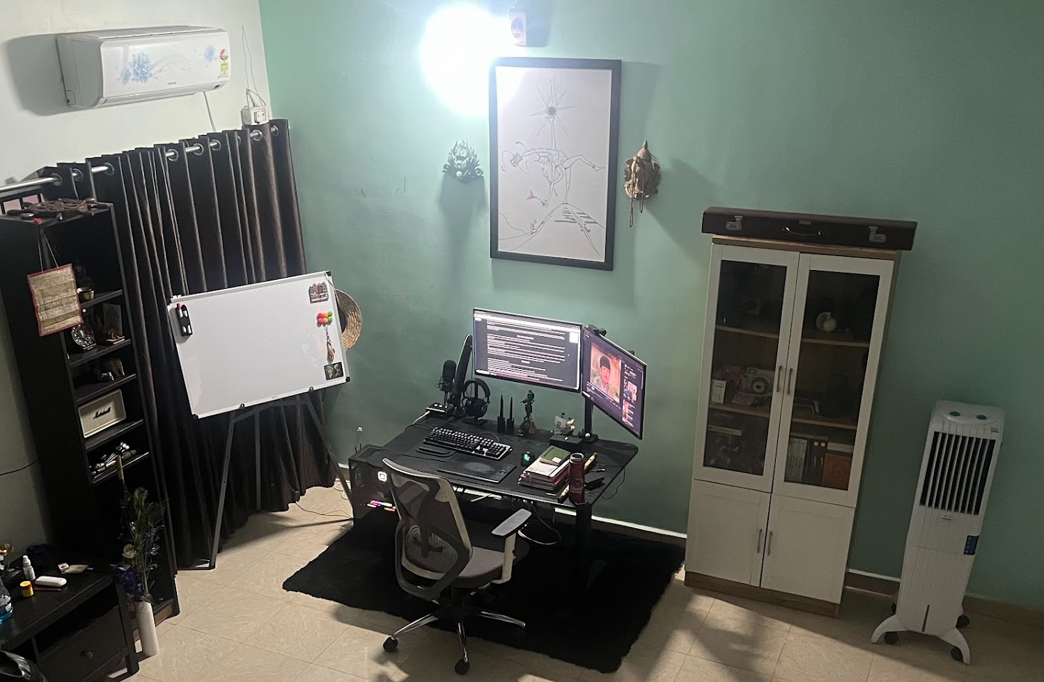Home office setup with a desk, dual monitors, keyboard, and mouse. Whiteboard on stand, black curtains, shelves with books and decor, glass-door cabinet, and a portable air cooler in a room with light green walls.