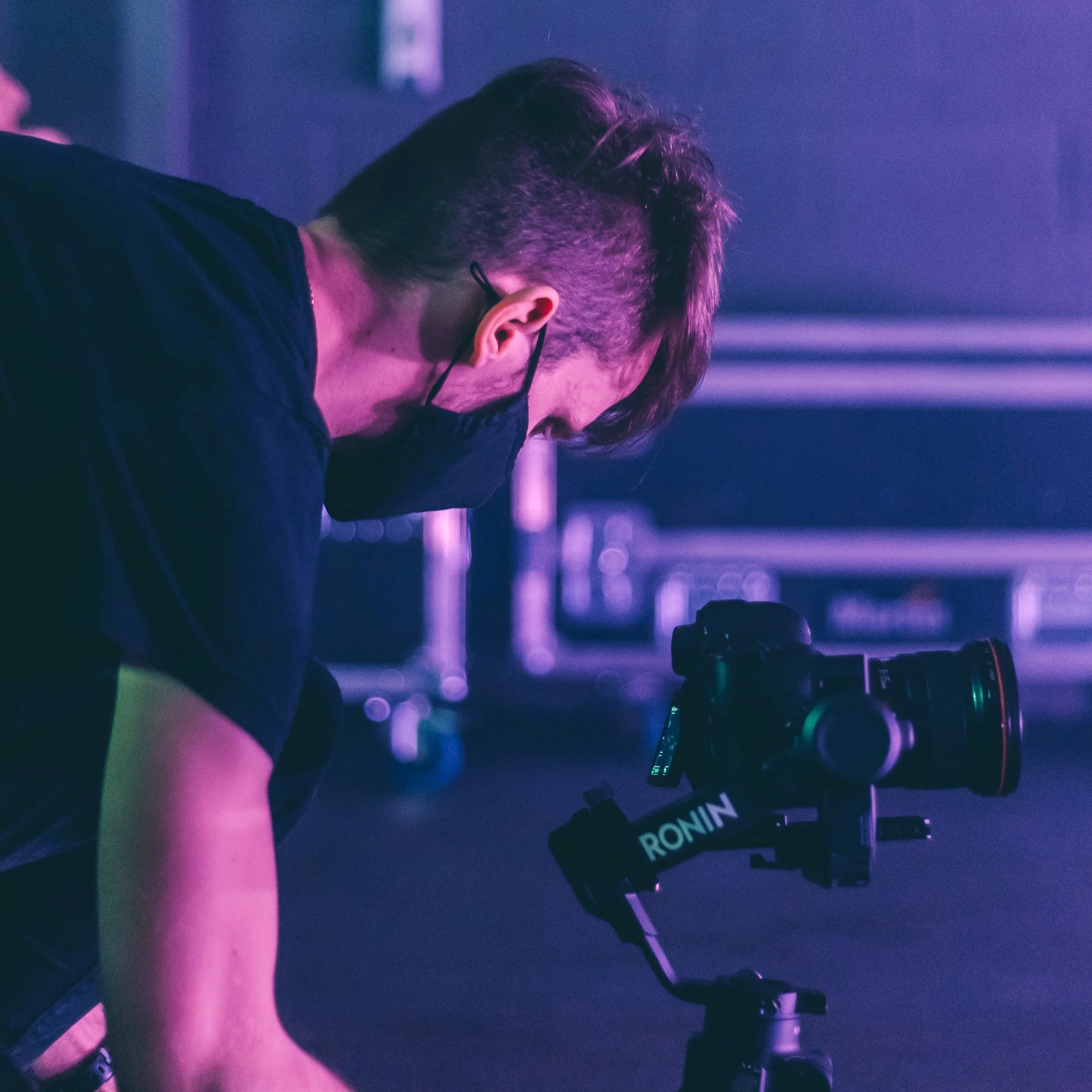 Photographer wearing a black mask and black shirt, using a Ronin stabilizer for a camera in a dimly lit setting with purple lighting.