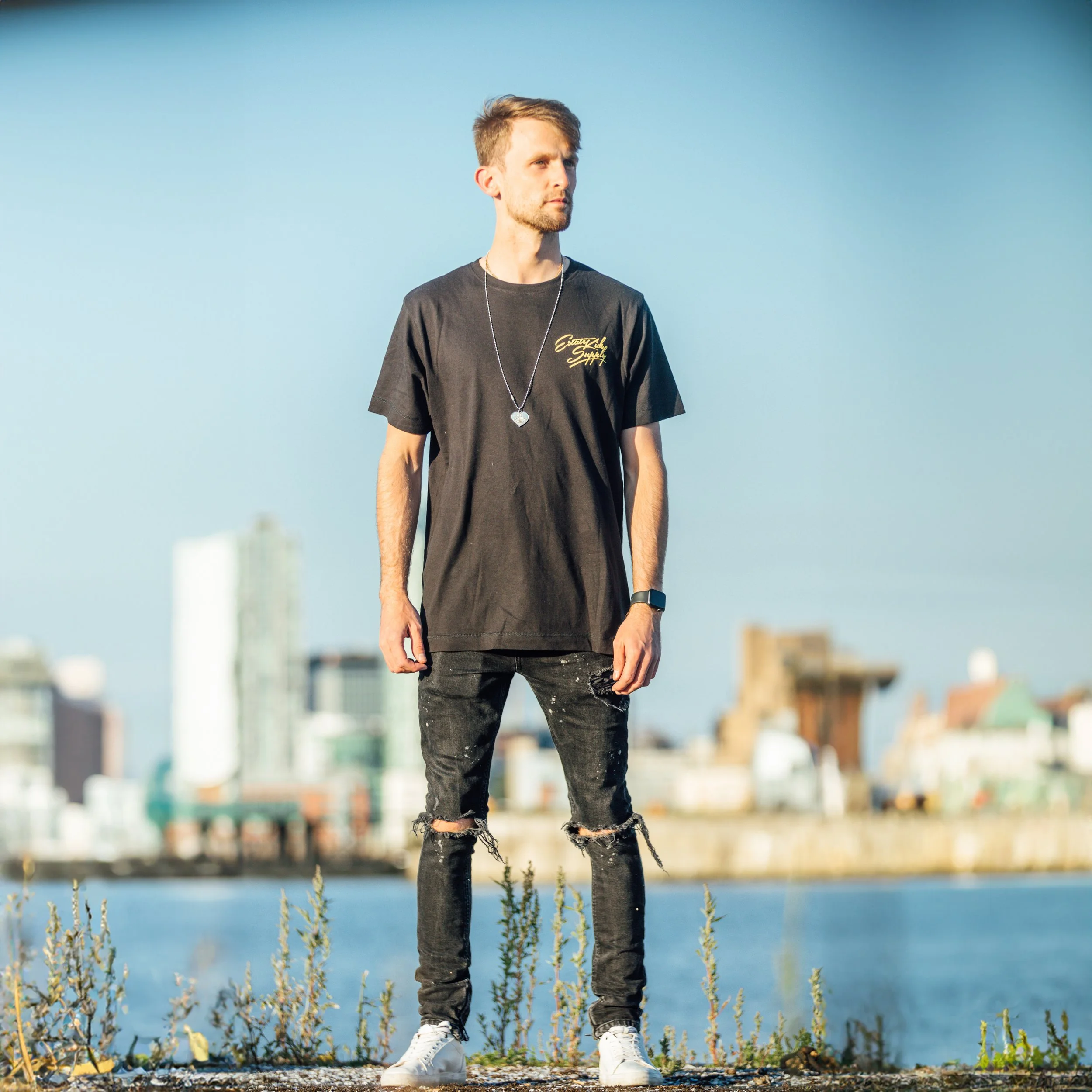A young man with short blonde hair stands outdoors by a body of water with a city skyline in the background. He's wearing a black t-shirt with yellow text, ripped black jeans, white sneakers, a silver necklace with a heart pendant, and a black smartwatch.