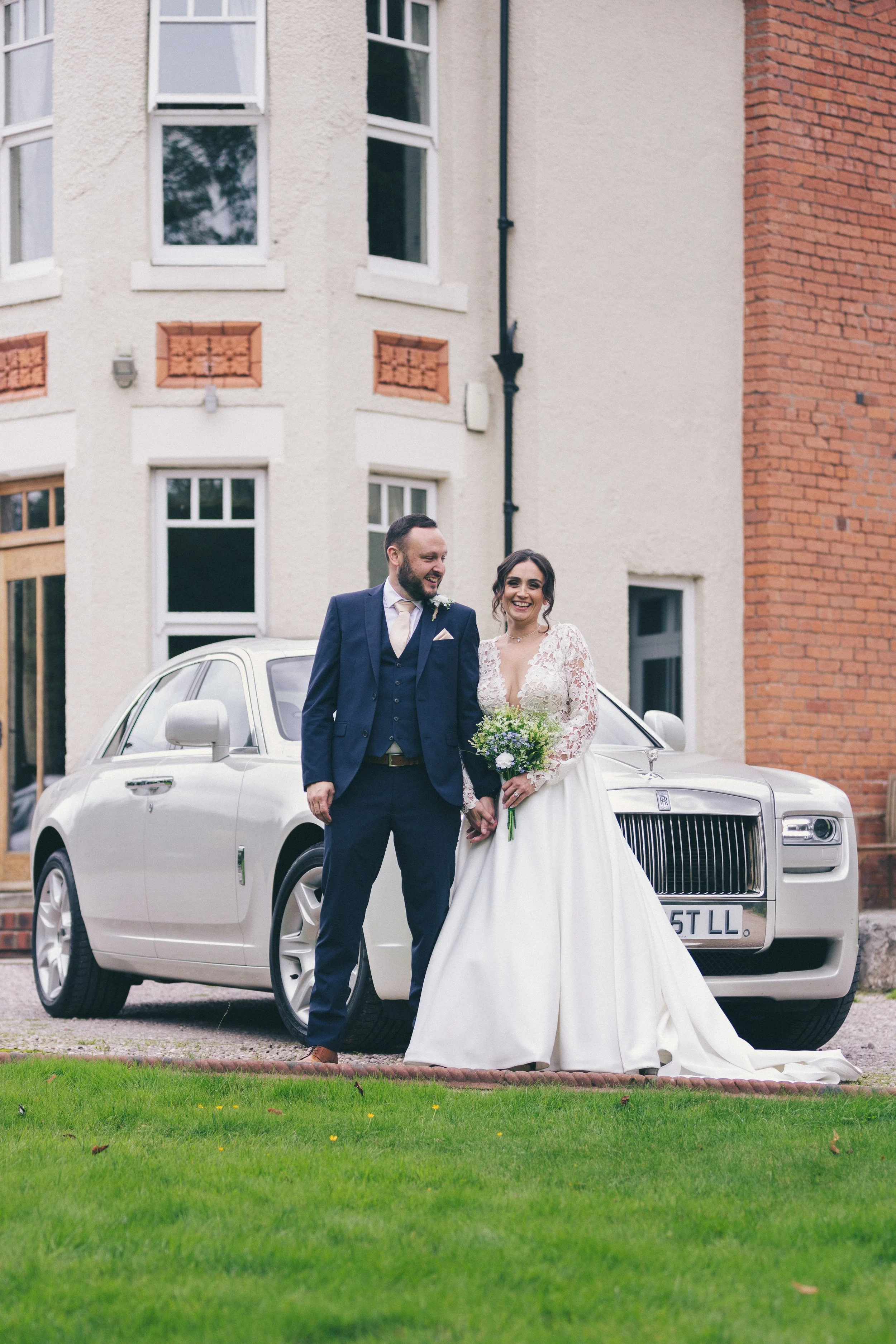 A newlywed couple in wedding attire standing in front of a white luxury car, smiling and holding hands, outside a building with white and red brick walls.
