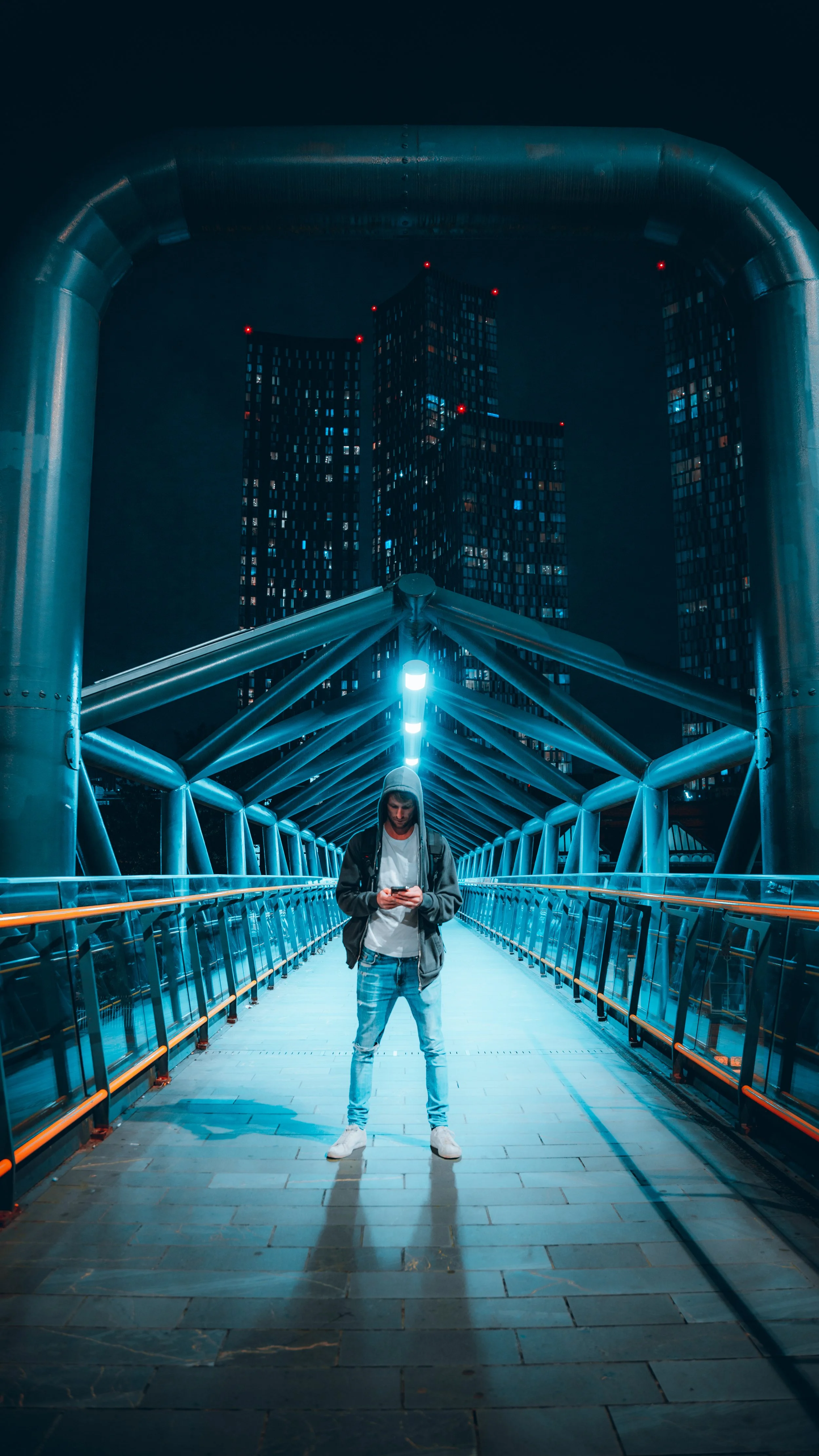 A man standing on a modern illuminated bridge at night, looking at his phone with city skyscrapers in the background.