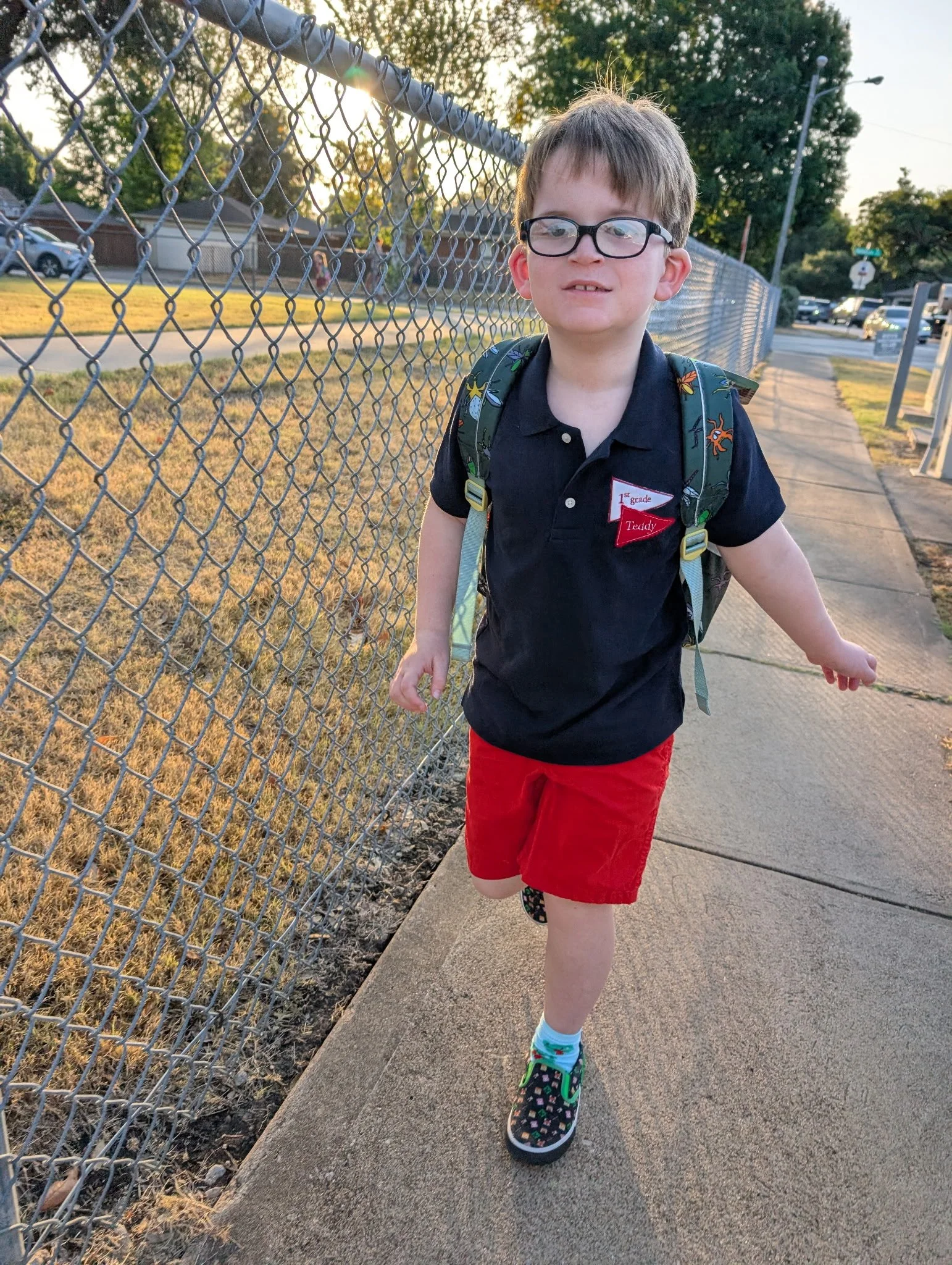 Child wearing a black shirt, red shorts, and a backpack while walking along a sidewalk.
