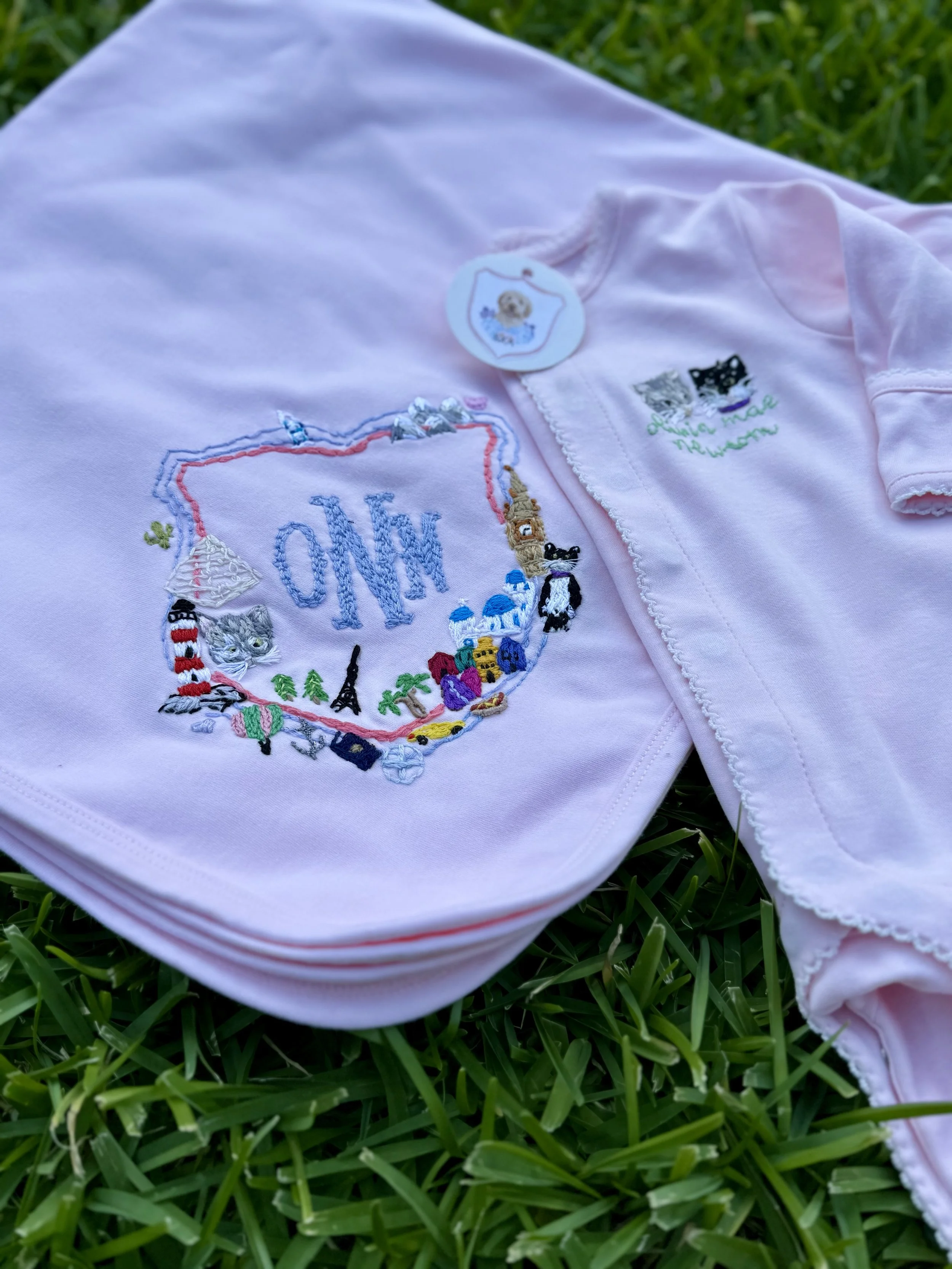 Light pink baby garment with colorful embroidered Paris-themed design, including landmarks, Eiffel Tower, and animals, laying on green grass.
