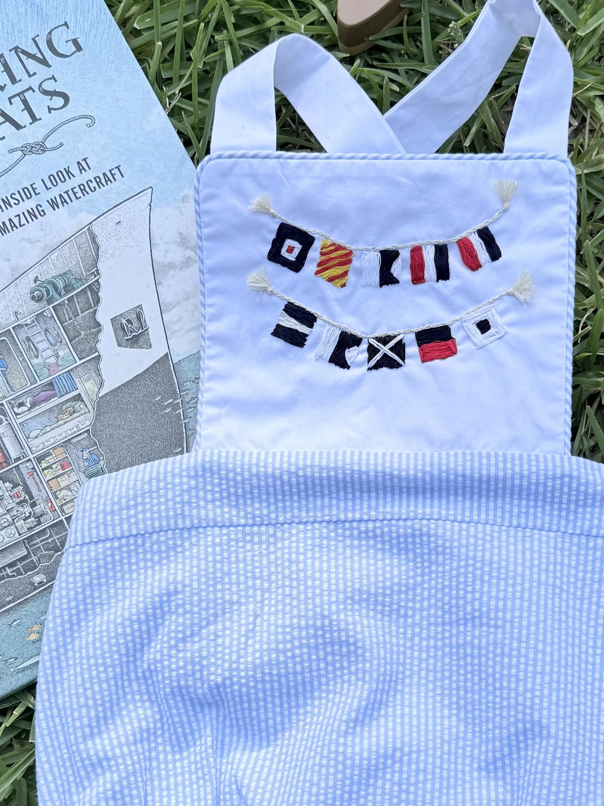 A white embroidered tote bag with navy, black, red, and yellow nautical signal flags on the front. The bag is laid on green grass next to a blue pamphlet titled "INSIDE LOOK AT AMAZING WATERCRAFT."