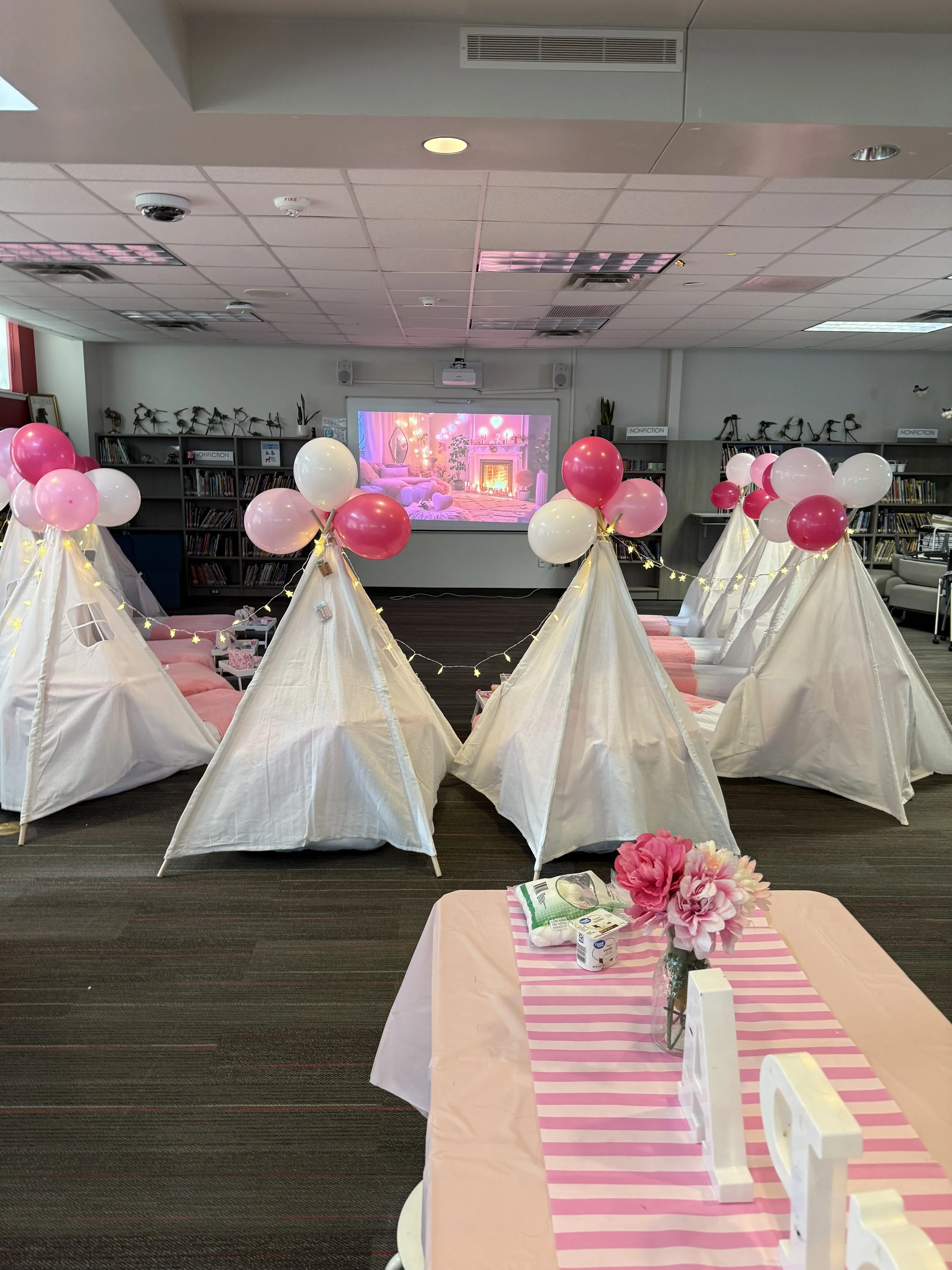 Pink-themed sleepover celebration with small white tents, pink and white balloons, and a striped table decorated with flowers.