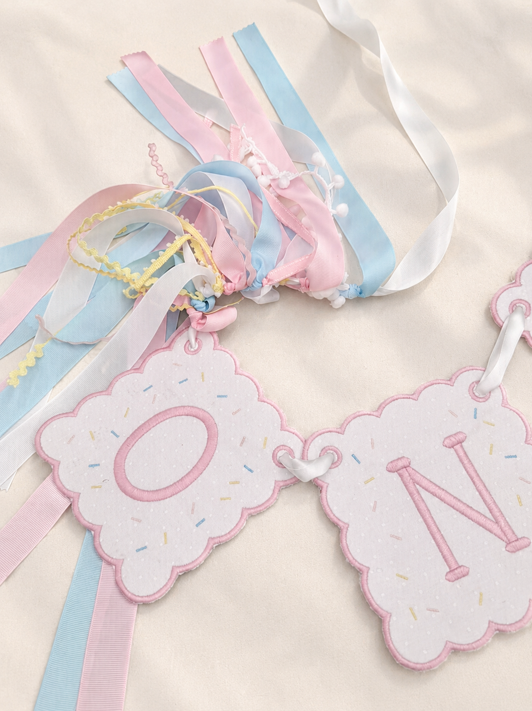Pastel embroidered birthday banner with scalloped pink pennants and pink, blue, white, and yellow ribbons.
