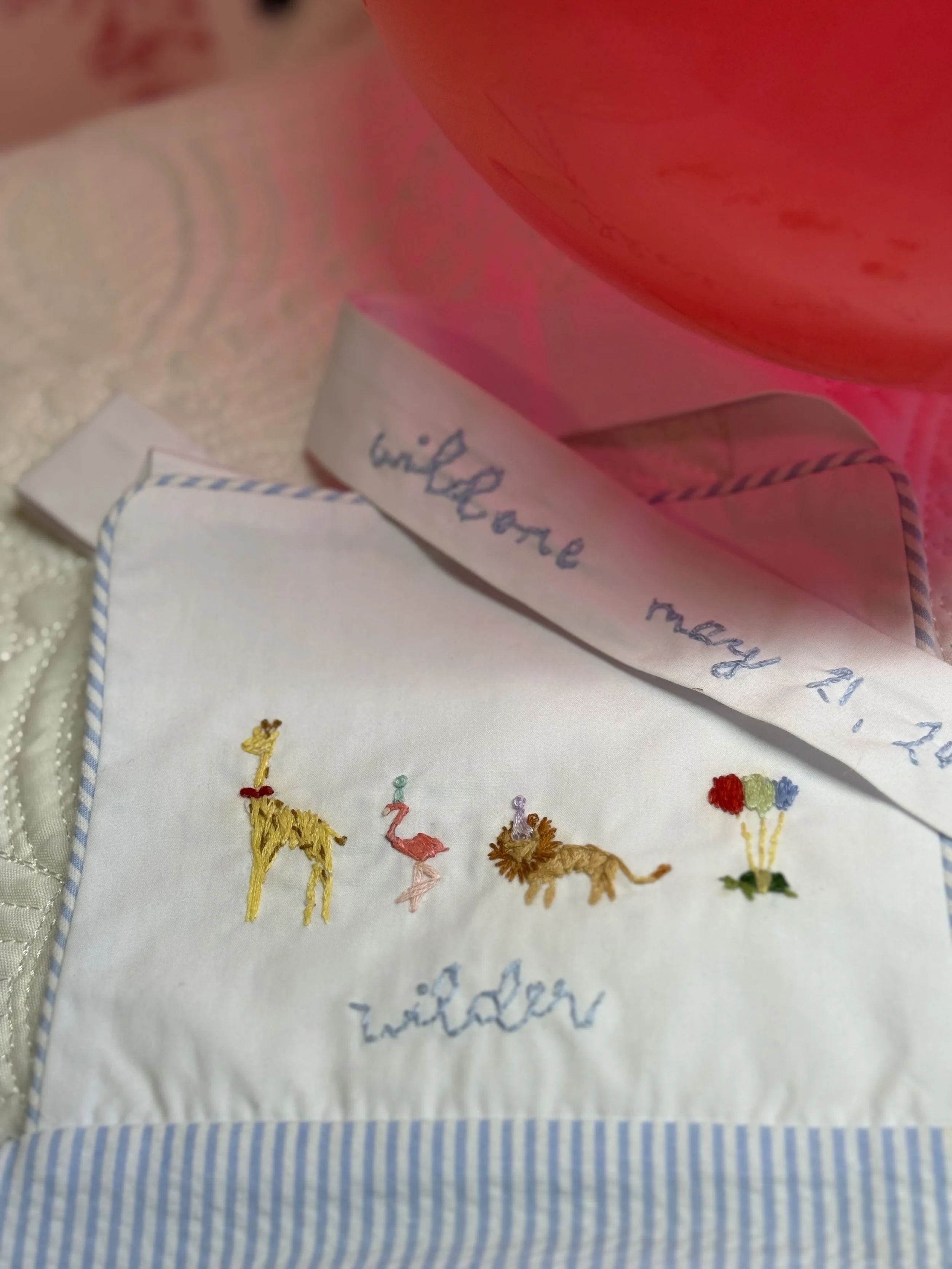 Embroidered fabric with animals and the words "wild" and "wilder" on white cloth, a label with handwritten note, and a red balloon in the background.