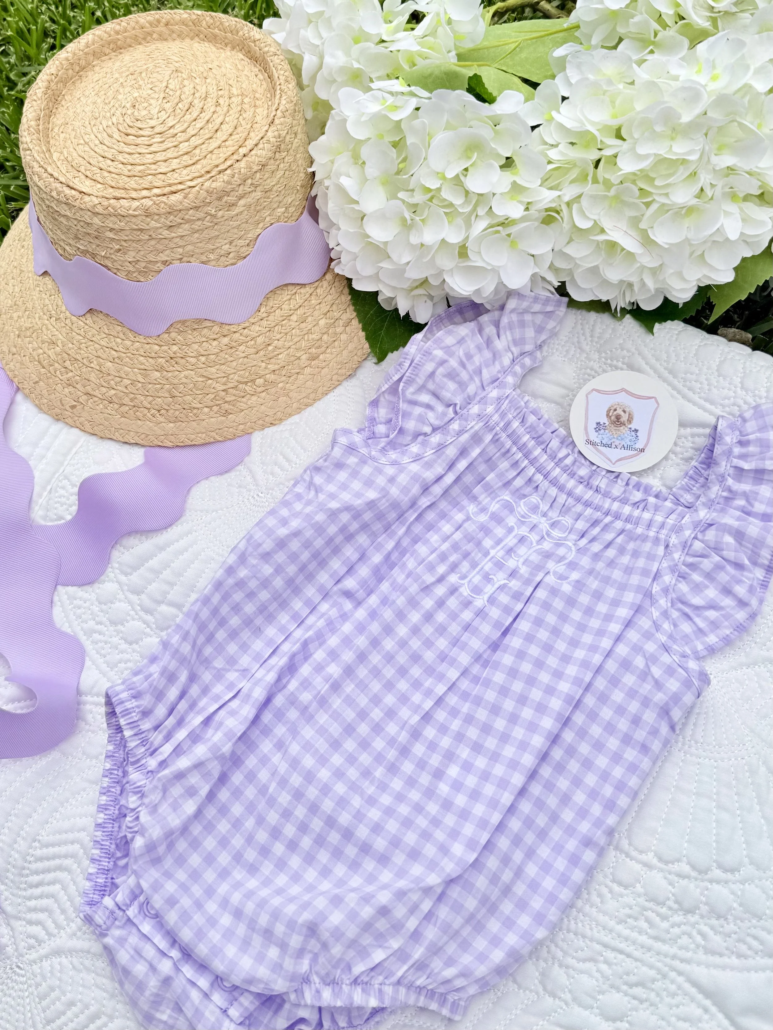 Lavender gingham children’s outfit beside a straw sun hat with lavender ribbon and white hydrangeas.