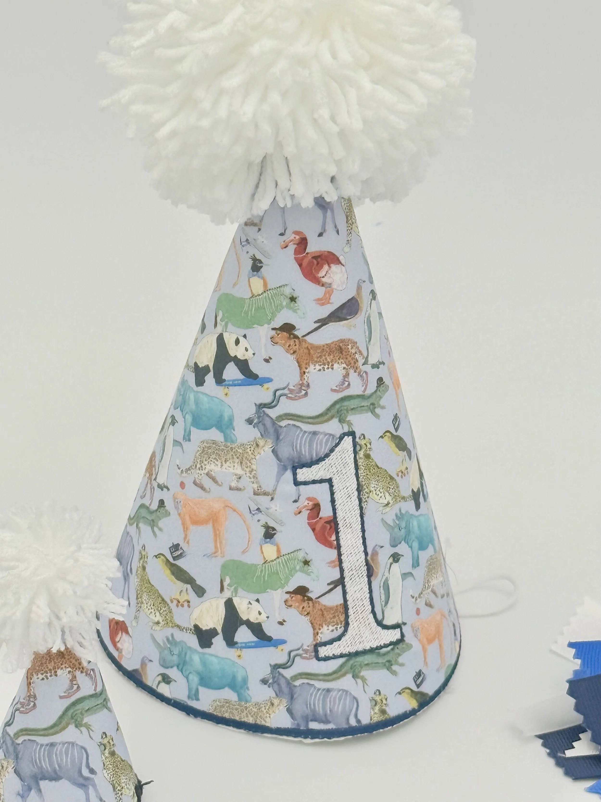 First birthday party hat with a colorful animal print, a large number 1, and a fluffy white pom-pom.