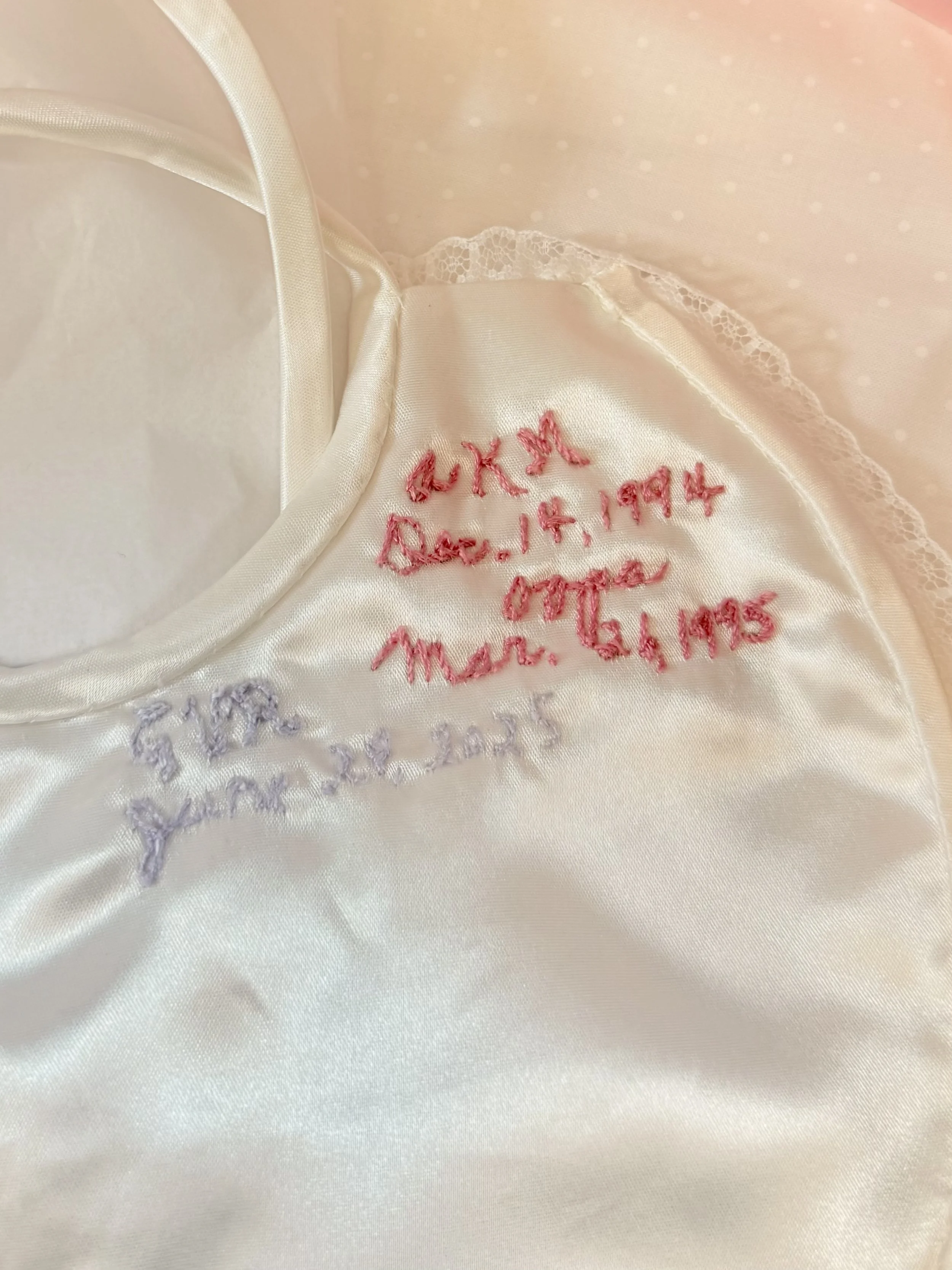 Close-up of a satin fabric with hand-stitched embroidery in pink and purple, including dates and names.