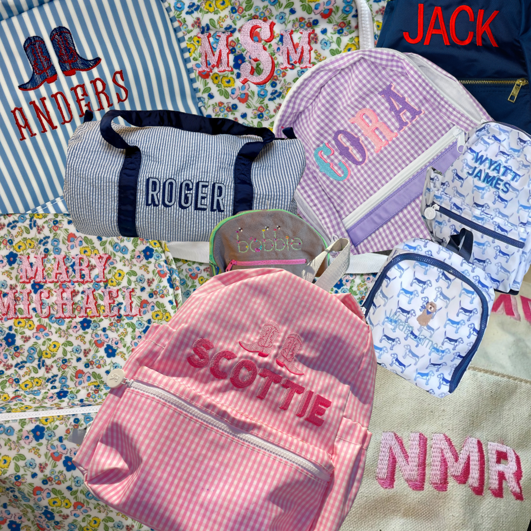 A collection of personalized children's backpacks, bags, and accessories with various colorful patterns and embroidery, some with names like Roger, Scoutie, Jack, Sophia, Wyatt James, and Mary McKean, arranged on a floral fabric background.