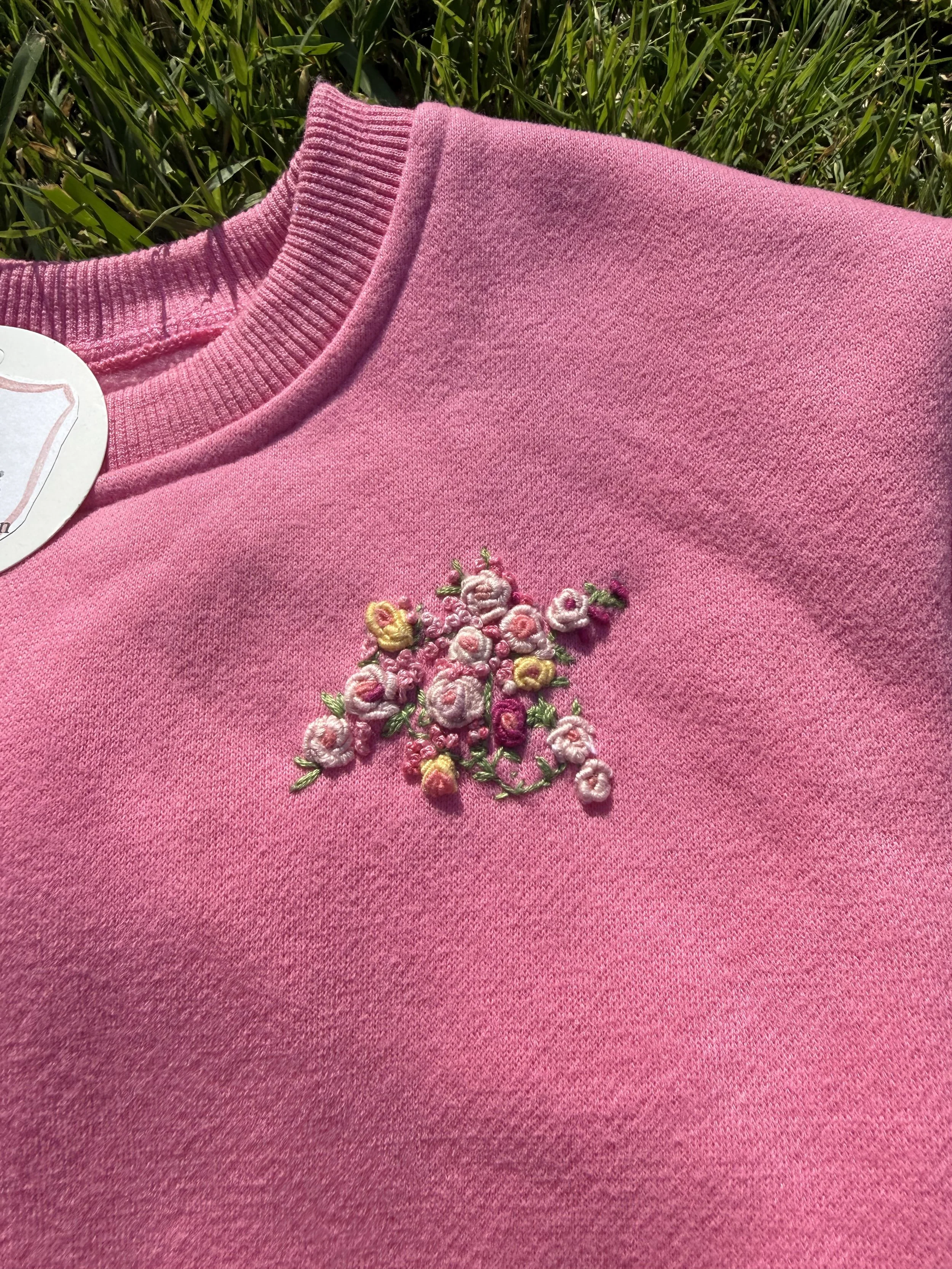 Close-up of a pink sweater with floral embroidery, lying on green grass.