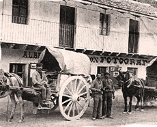 Photo ancienne montrant une charrette tirée par un cheval devant un bâtiment. Des hommes se tiennent près de la charrette. Le bâtiment a une inscription indiquant "Fotografía".