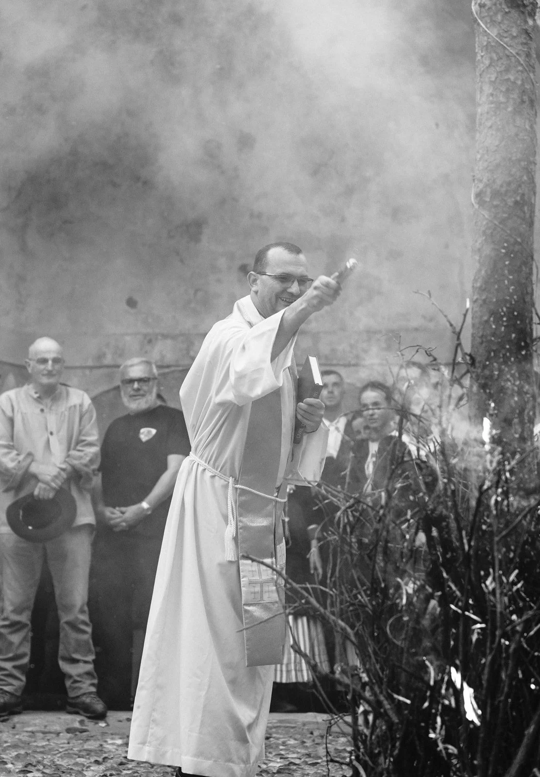 Un prêtre en robe blanche bénit un feu en extérieur, entouré par des spectateurs.